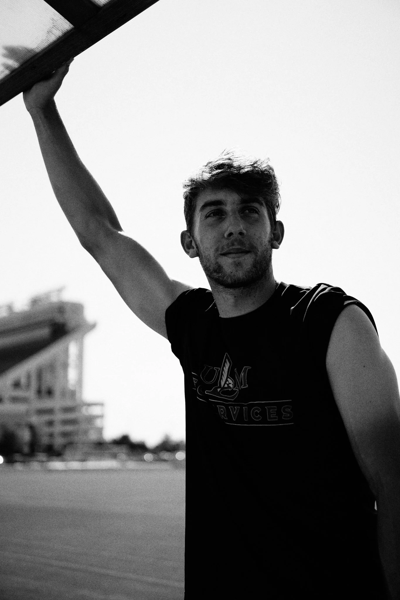 A young man with tousled hair and a sleeveless T-shirt, holding a skateboard overhead, standing outdoors with a building and a minimalist urban background.