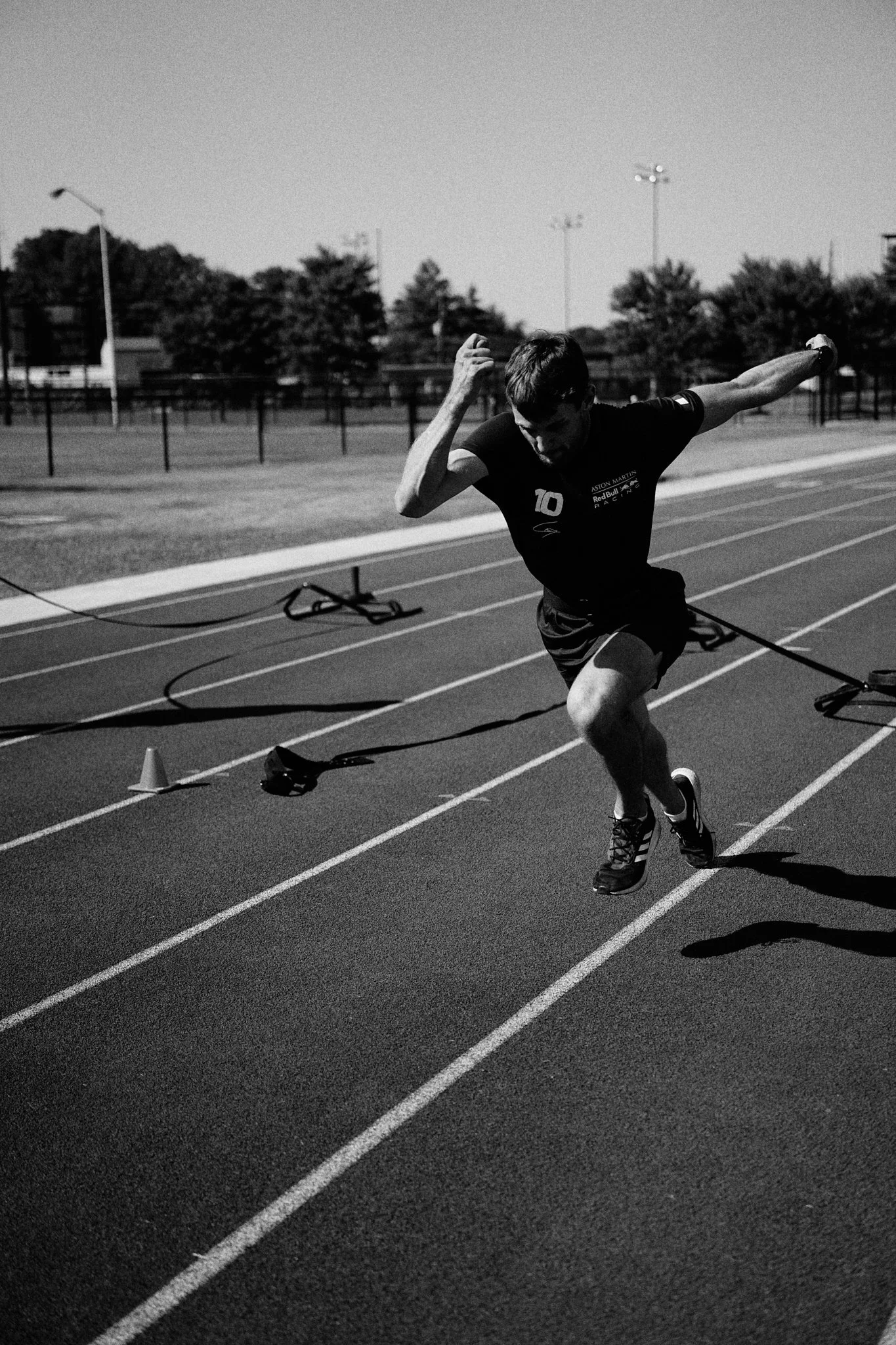 Black and white photo of a male athlete sprinting on a running track, crossing the finish line, with poles, cones, and trees in the background.