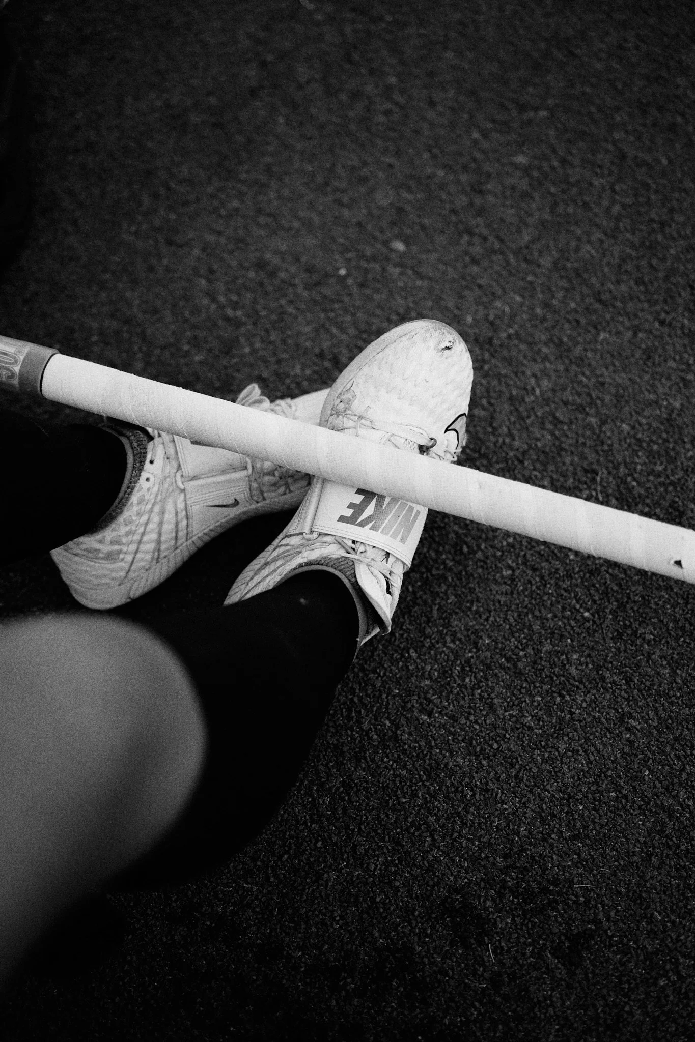 Black and white photo of a person wearing white Nike sneakers and black leggings, balancing a hockey stick on their shoes on a textured sports court.