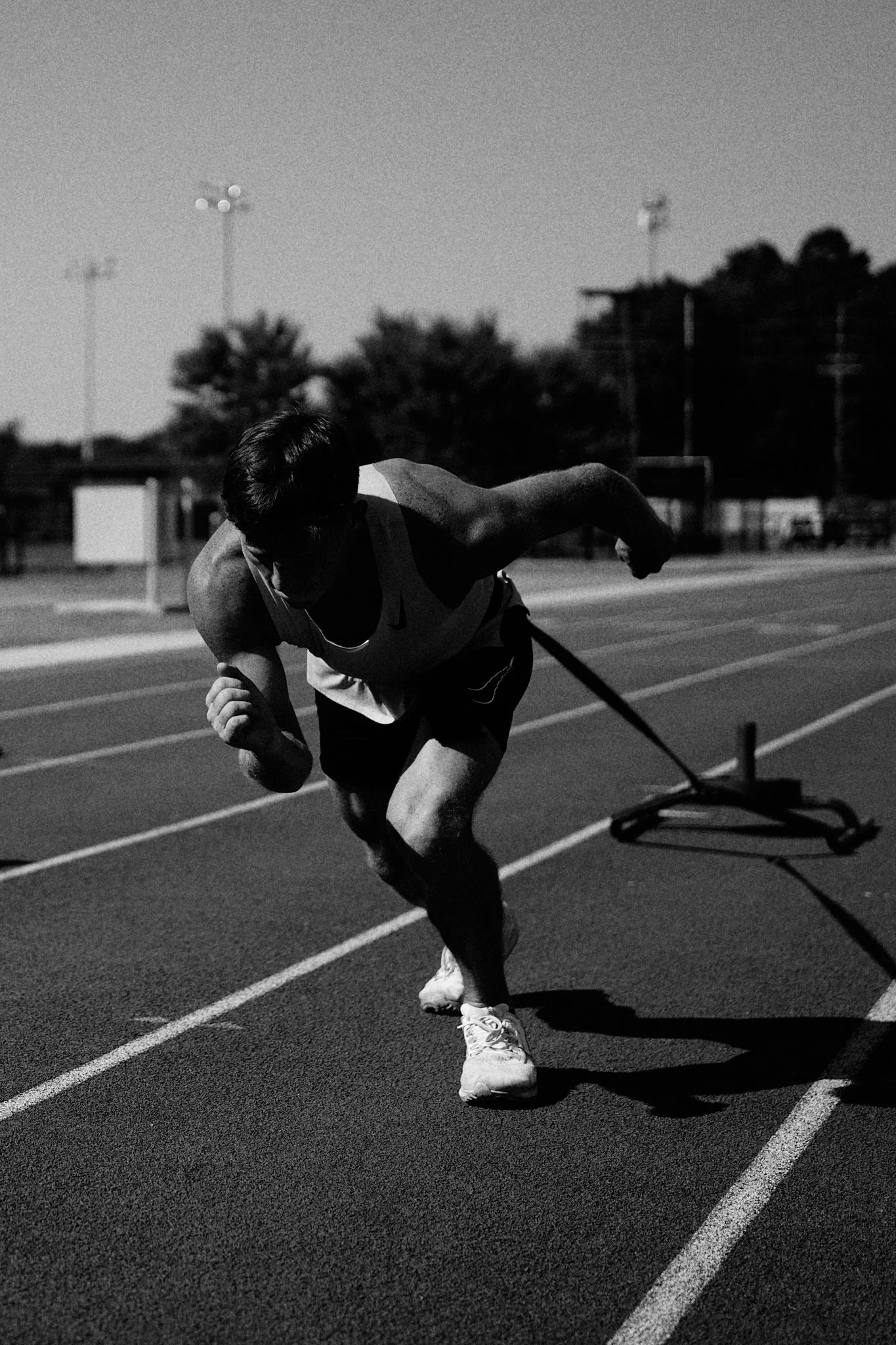 A male sprinter in athletic gear running on a track, captured in black and white, with sunlight casting shadows.