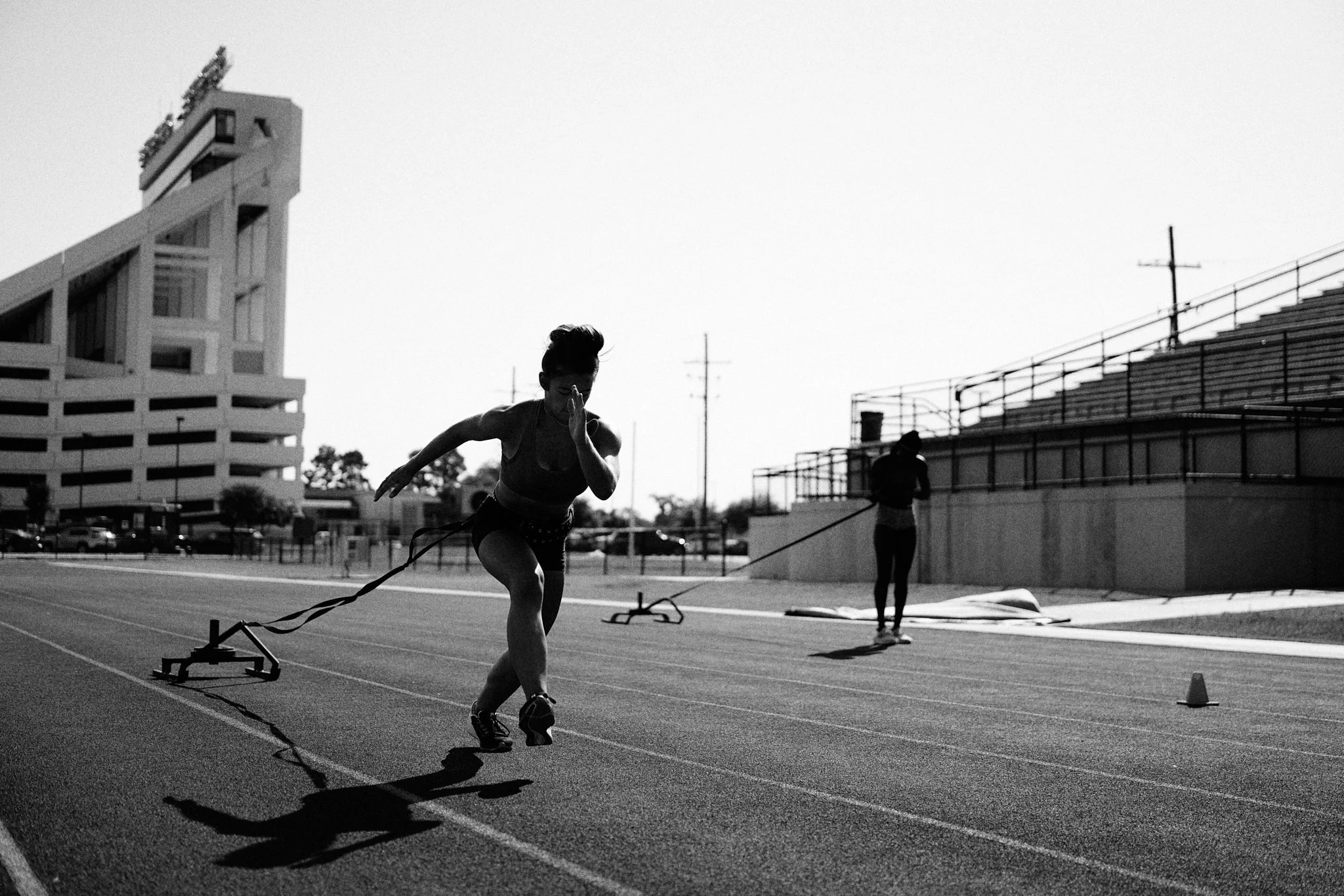 A woman sprinting on a track with a resistance sled attached, while another person stands in the background. The scene is in black and white, with a large building and empty bleachers behind them.