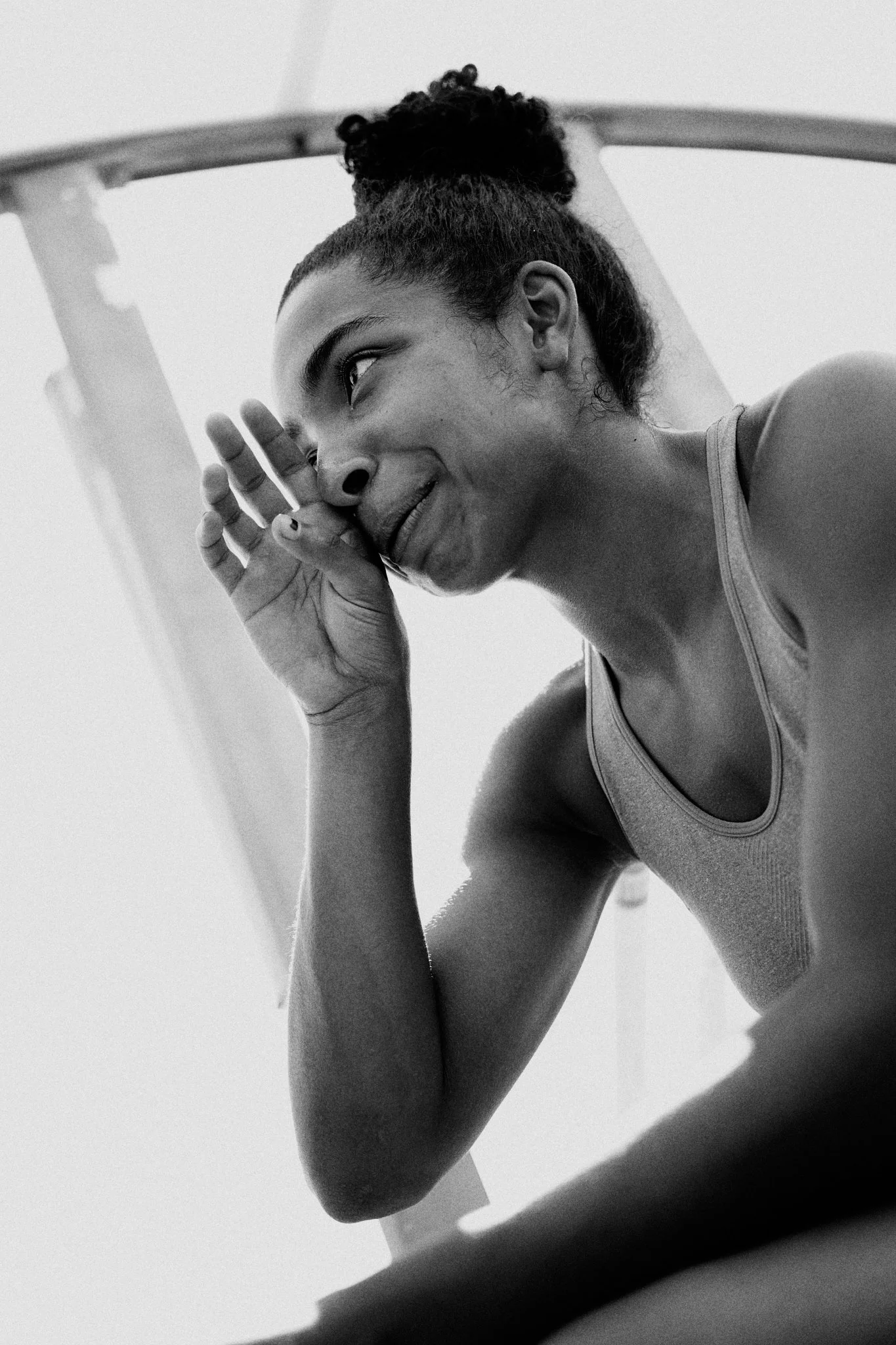 A young woman with curly hair in a bun, wearing a tank top, resting her head on her hand with a thoughtful expression.