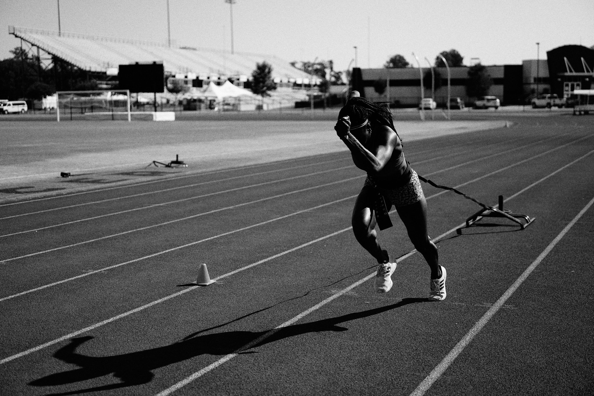 A female sprinter runs on an outdoor track, pulling a sled for resistance training, during daylight. She is in mid-stride, wearing athletic gear and running shoes, with her long hair flowing behind her.