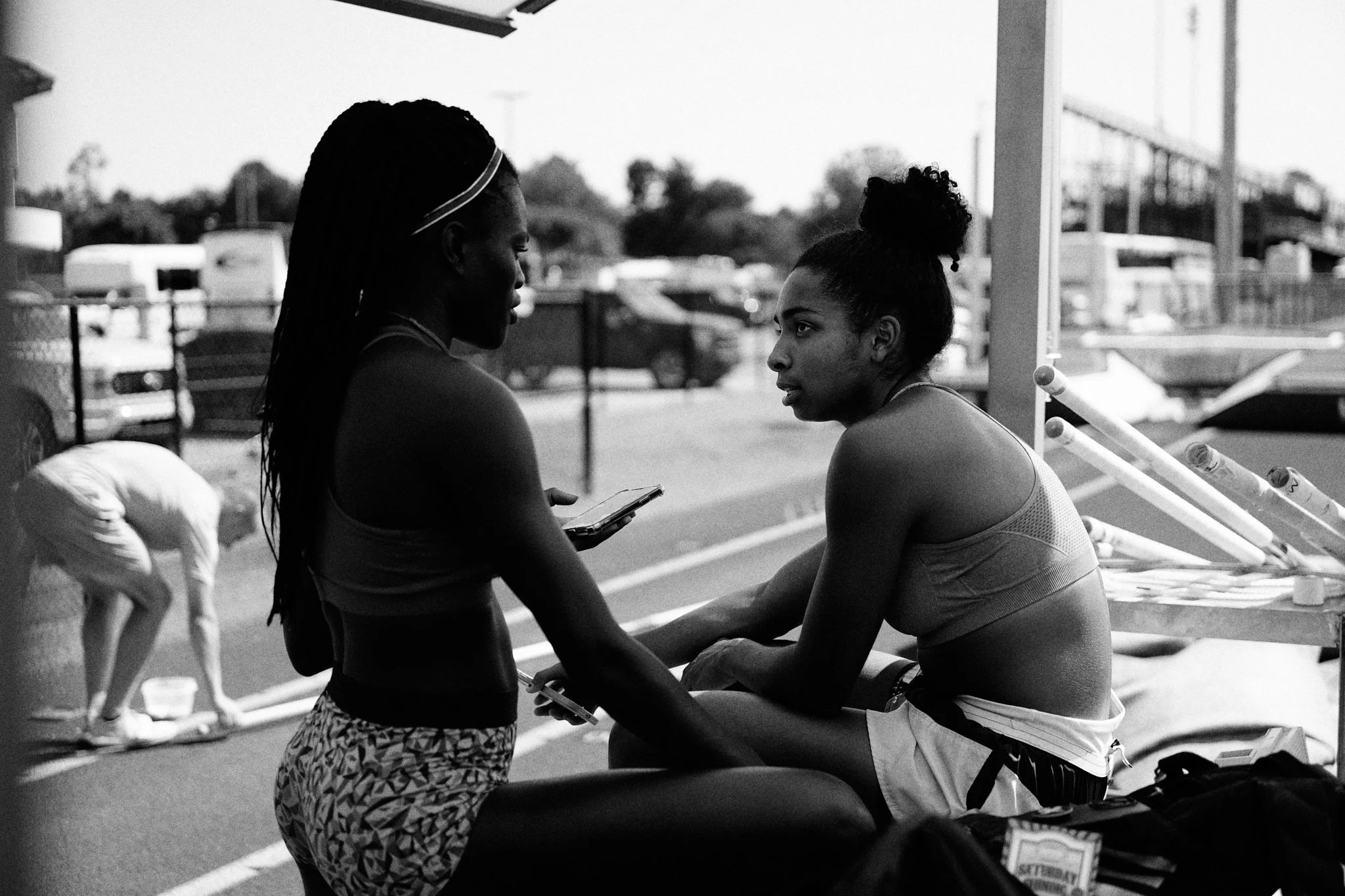 Two young women in athletic clothing having a conversation on a sports field, with one sitting and the other standing, and a person in the background bending down near a bench or equipment.