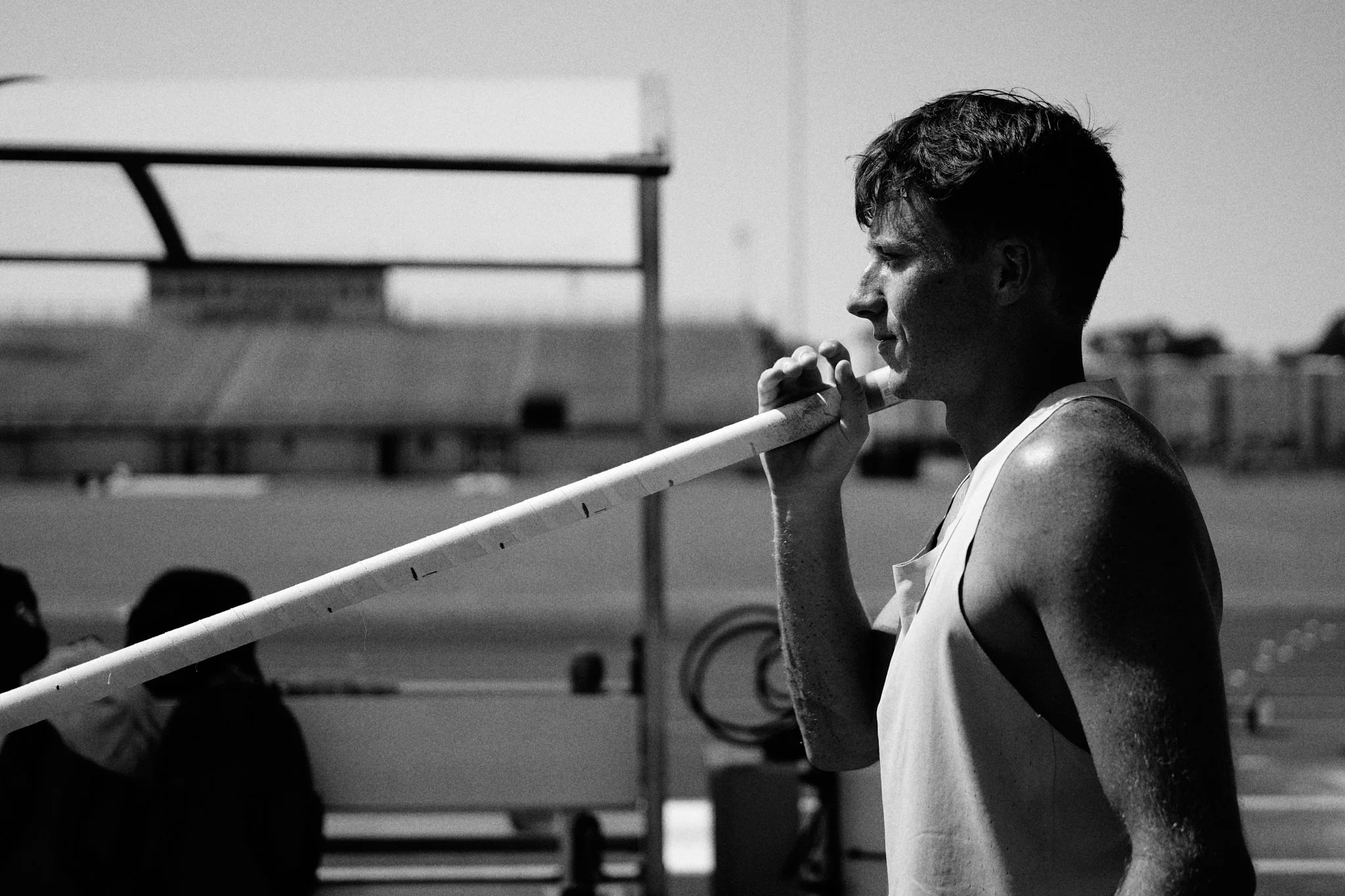 A young male athlete, in a sleeveless shirt, stands outdoors and rests a pole on his shoulder, looking thoughtful.