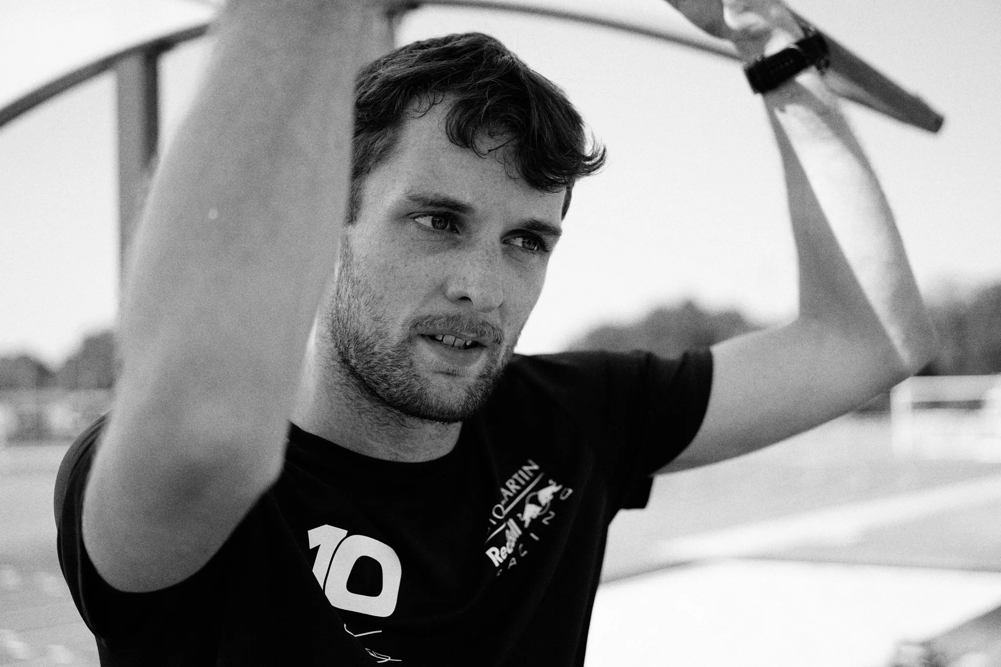 A young man with dark hair and a beard, wearing a black sports t-shirt, is holding a sports racket over his head, possibly preparing for a game or practice session on an outdoor sports court.