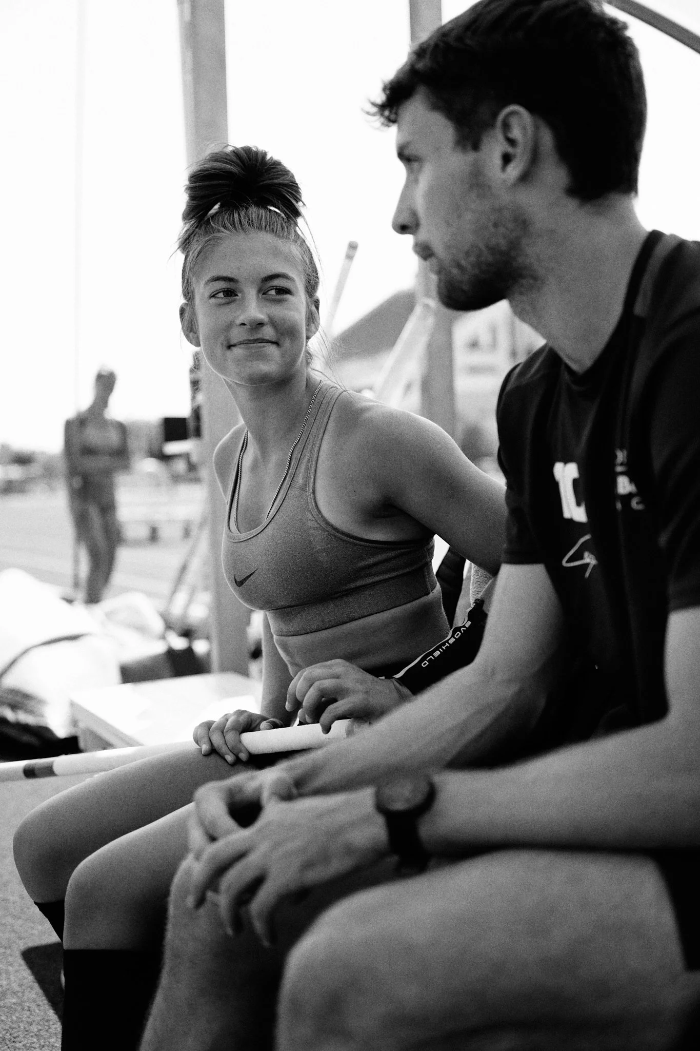 A black-and-white photo of a young female athlete sitting and smiling at a male coach or trainer, with a blurred person in the background and athletic equipment around.