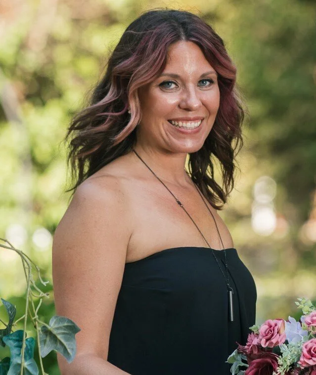 Smiling woman with wavy dark hair and pink highlights wearing a black strapless dress outdoors among flowers