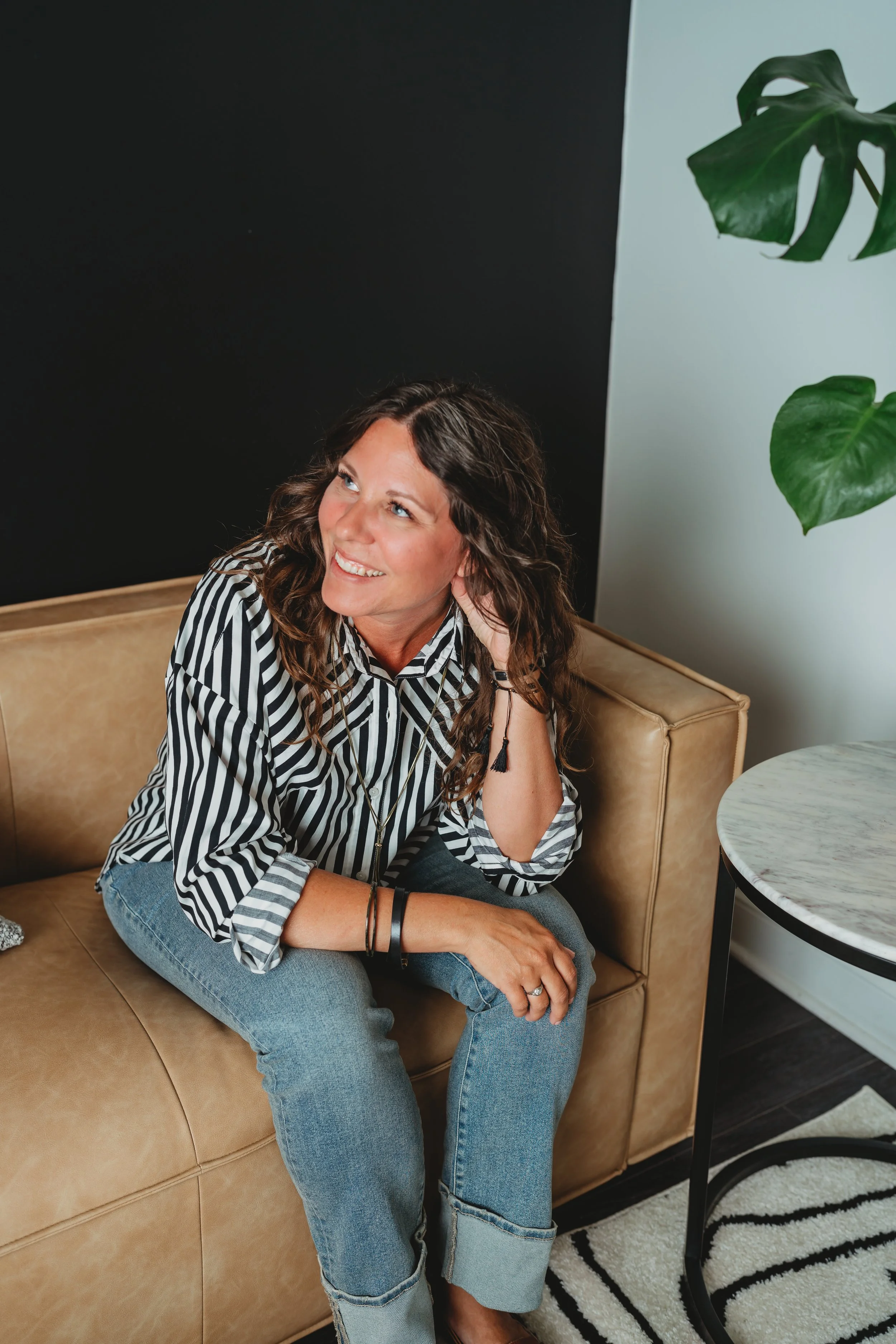 Smiling woman in a black-and-white striped blouse sitting on a tan sofa beside houseplants