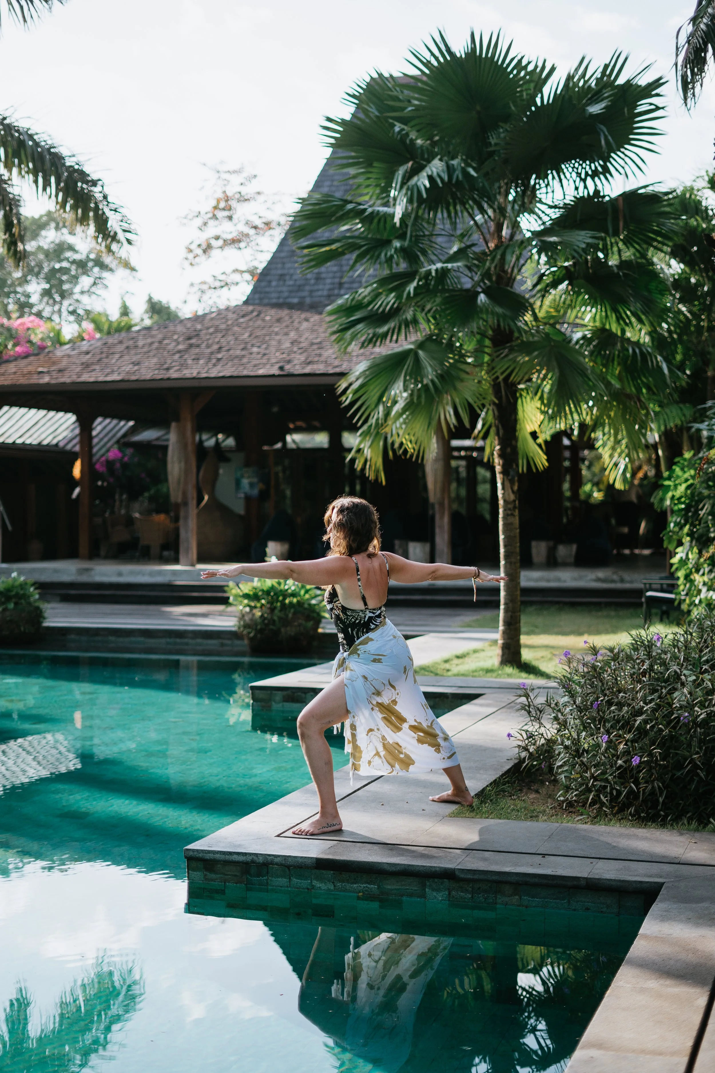 Woman practicing yoga beside a swimming pool at a tropical resort surrounded by palm trees