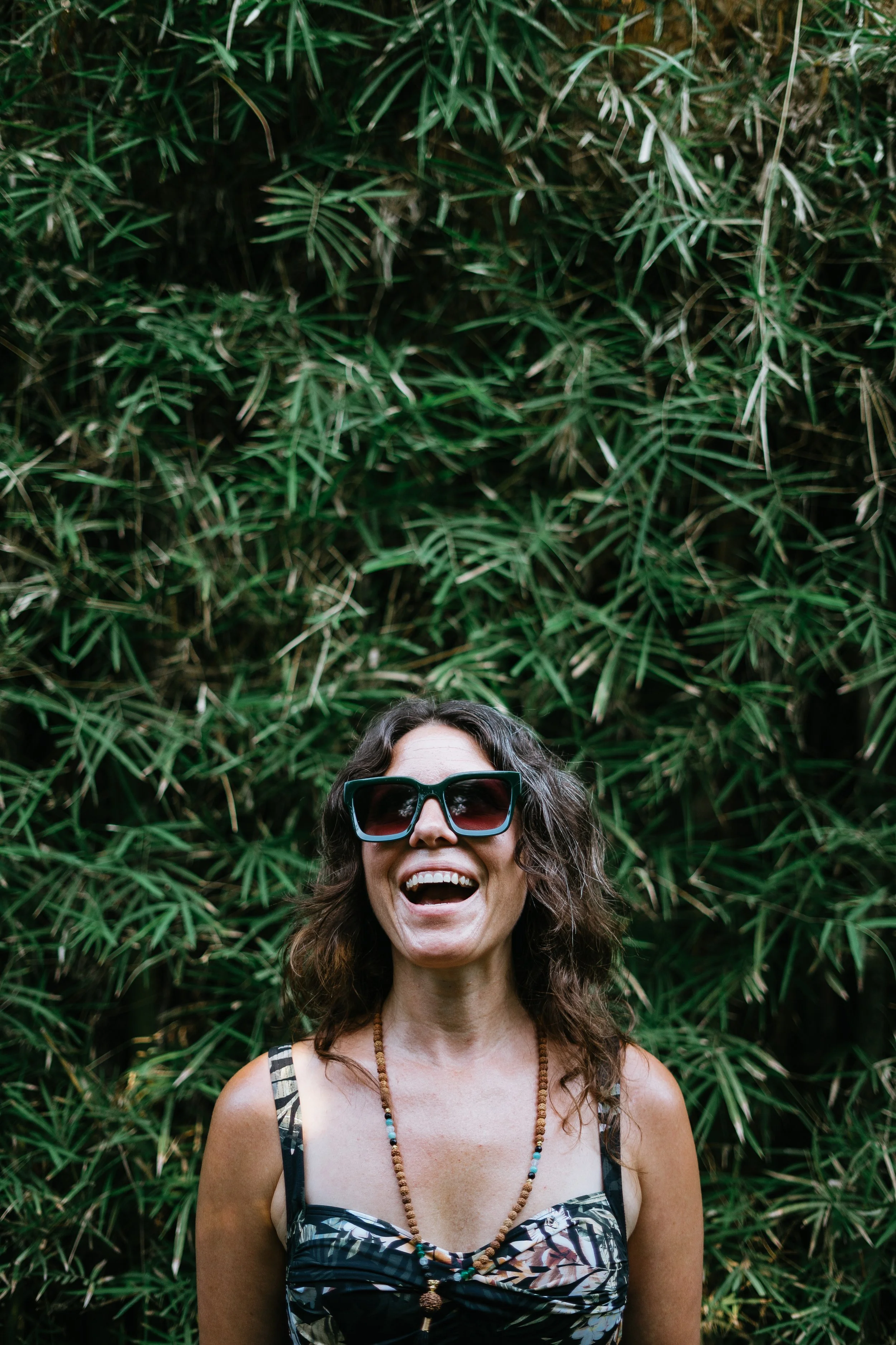 Smiling woman wearing oversized sunglasses and a beaded necklace in front of lush green foliage