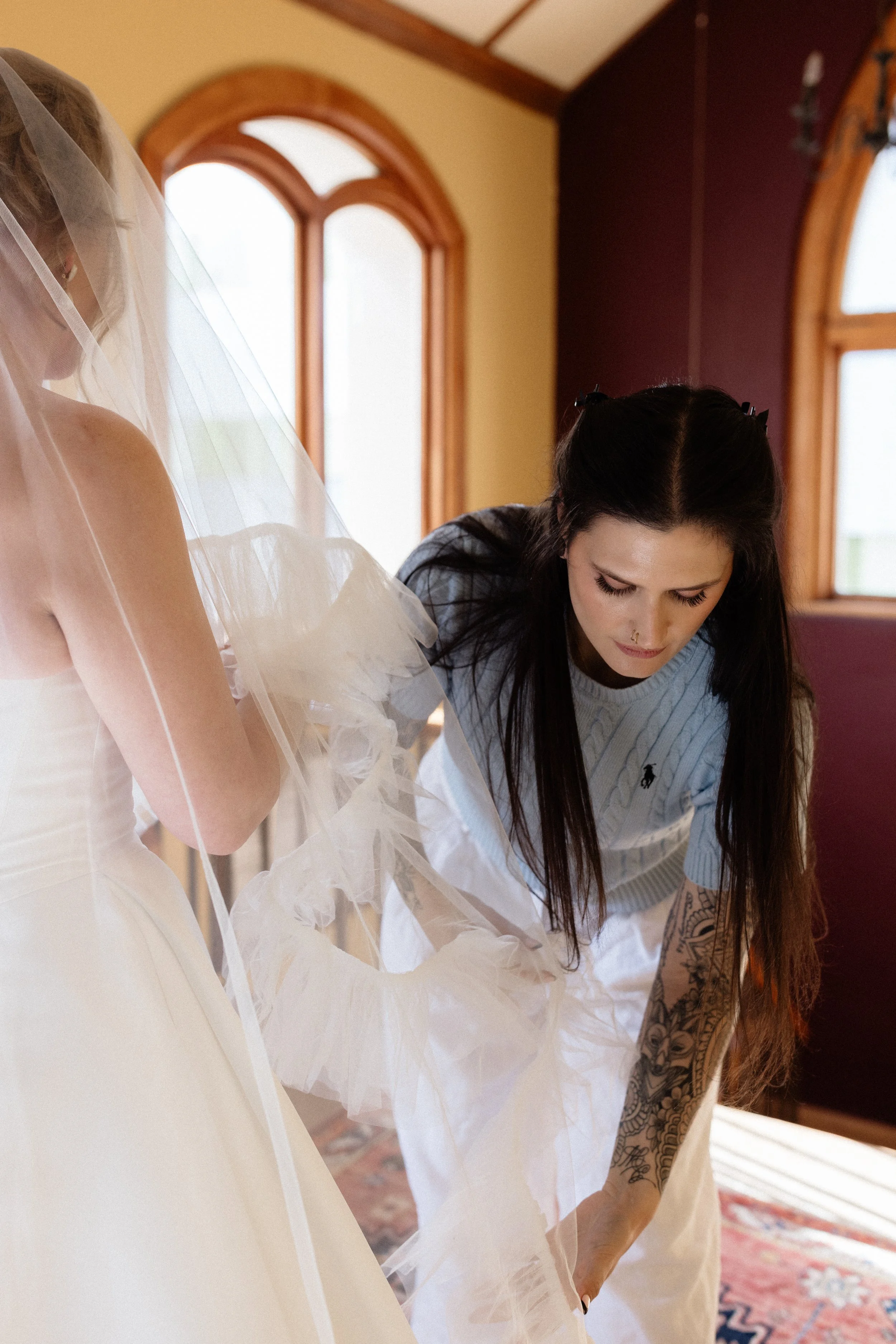 A woman in a blue shirt with tattoos on her arm is adjusting bride's wedding dress and veil in a room with large arched windows and a colorful rug.
makeup artist
hair artist
getting bride ready
destination wedding 
uk wedding
