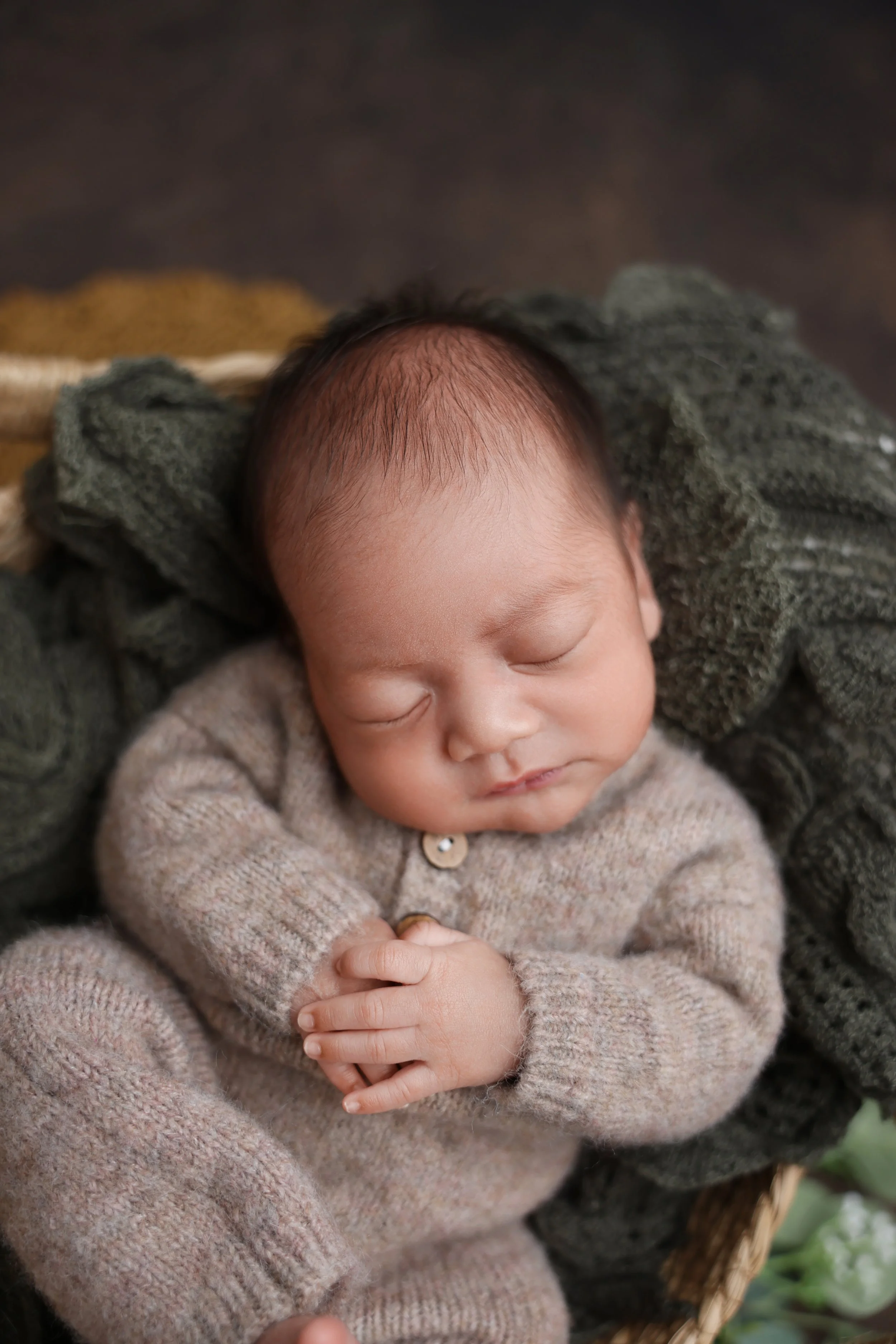 A sleeping newborn baby with dark hair, wearing a beige knitted onesie, curled up with hands clasped in front, nestled in a basket with soft green and beige blankets.