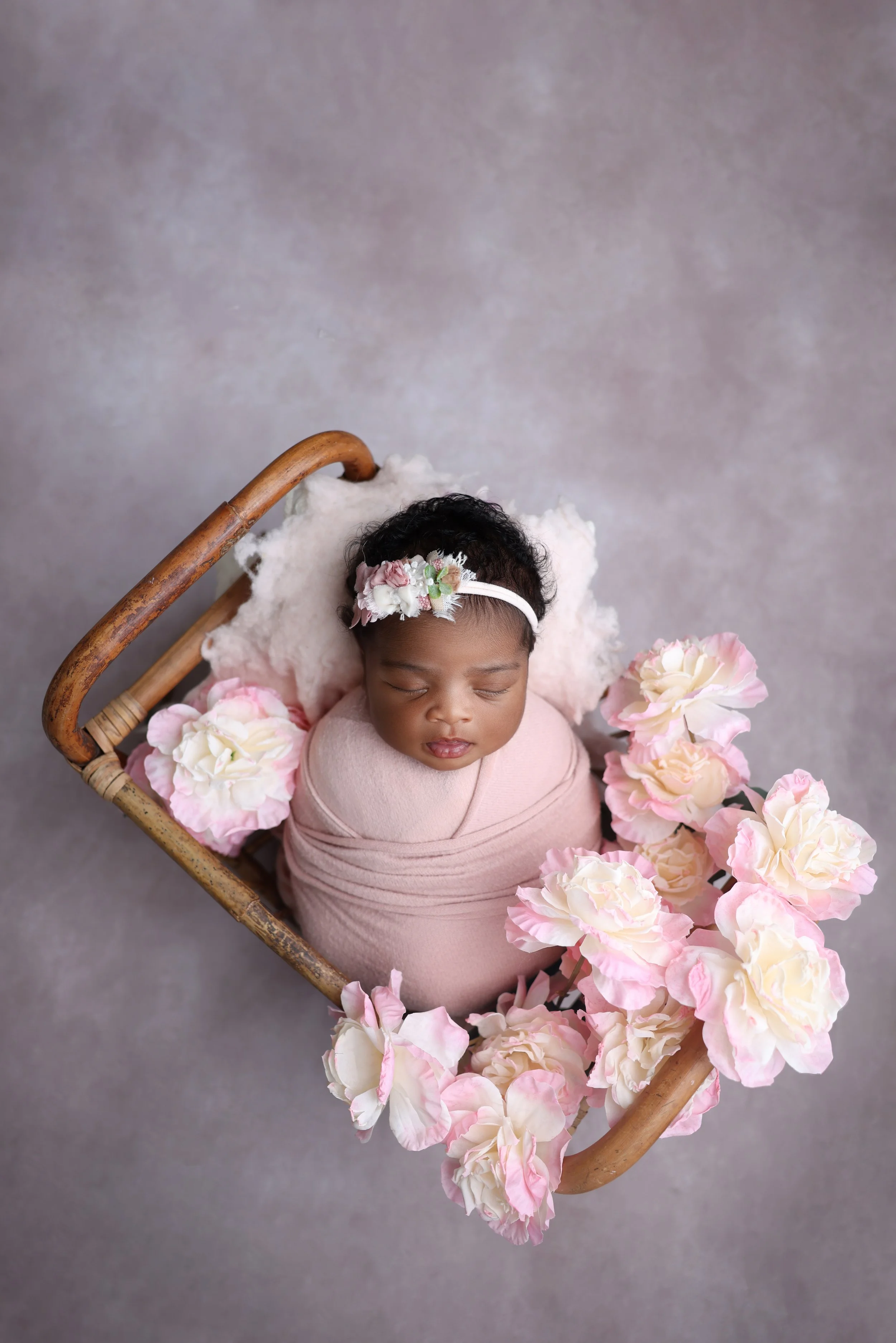 A sleeping baby girl wrapped in a pink cloth, lying in a small wooden basket adorned with pink and white flowers, with a floral headband on her head, against a soft gray background.
