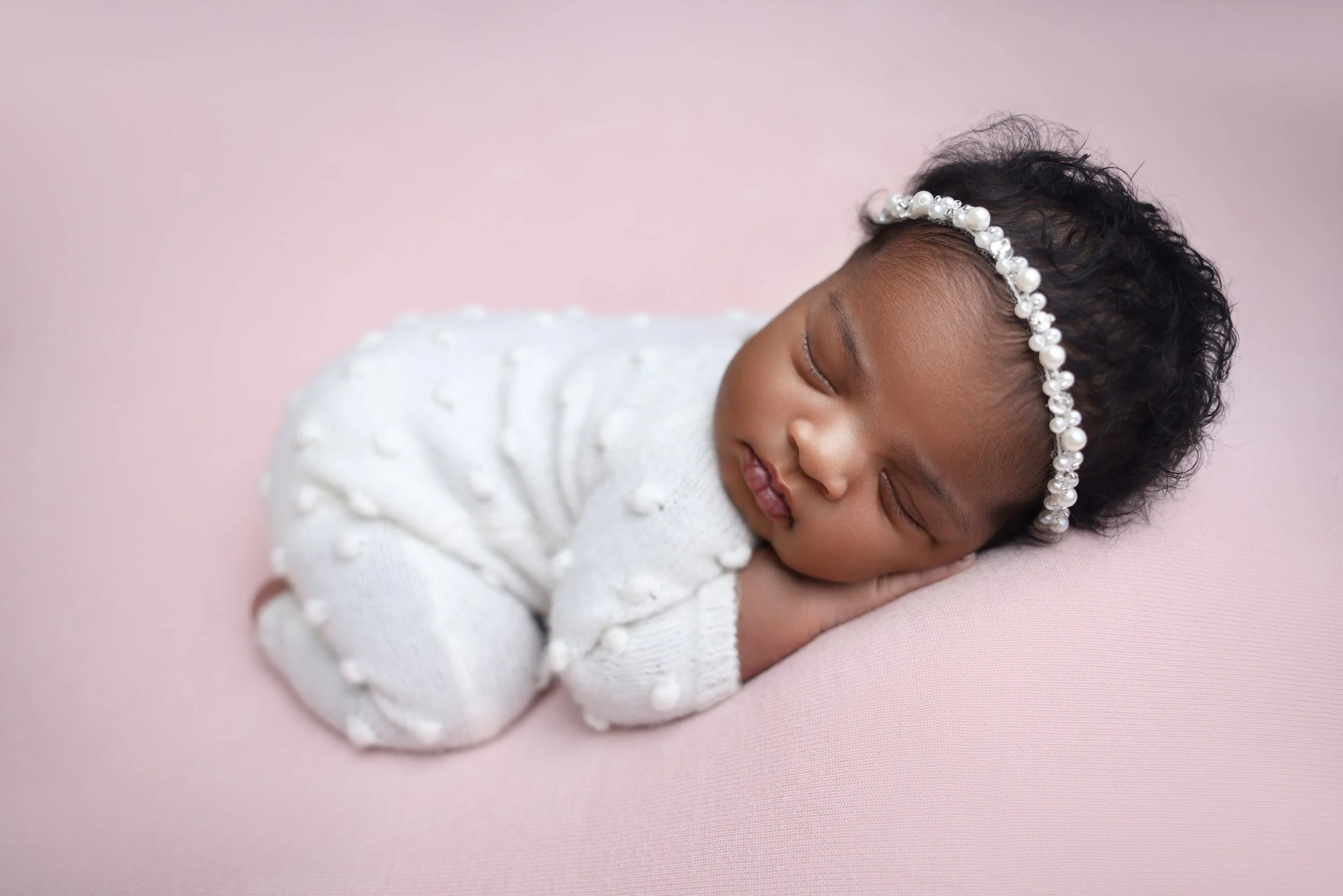 A sleeping baby girl with dark, curly hair, wearing a white headband with pearls, lying on a pink surface, dressed in a white sweater with textured dots.