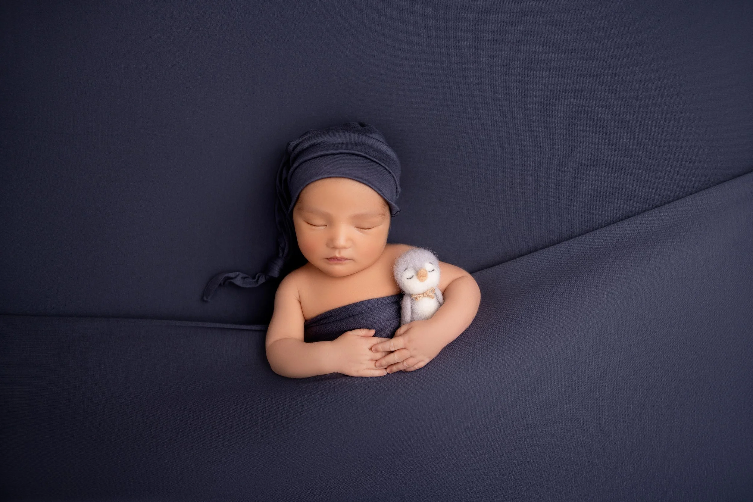 Baby sleeping on a dark blue fabric surface, holding a small stuffed penguin toy, with a matching dark blue hat and wrap.
