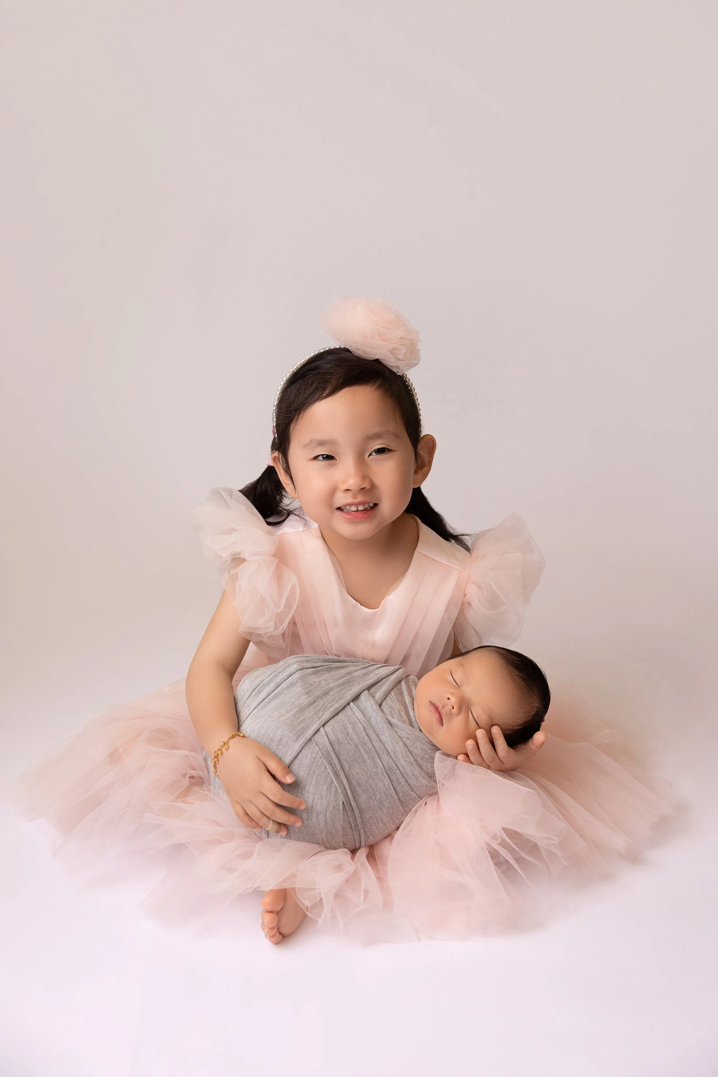 A young girl in a pink dress with tulle says hello to her newborn sibling, who is wrapped in a gray blanket and sleeping peacefully.