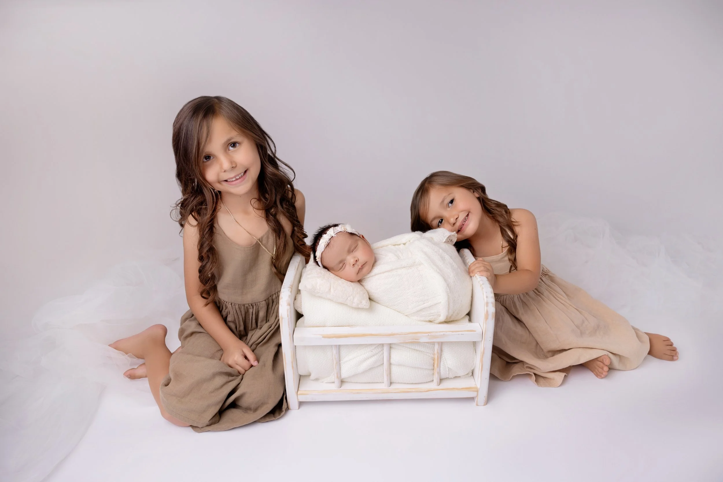 Three young girls, two older and one baby, with the baby sleeping in a white crib and the older girls kneeling next to it, all dressed in beige and tan clothing, on a white background.