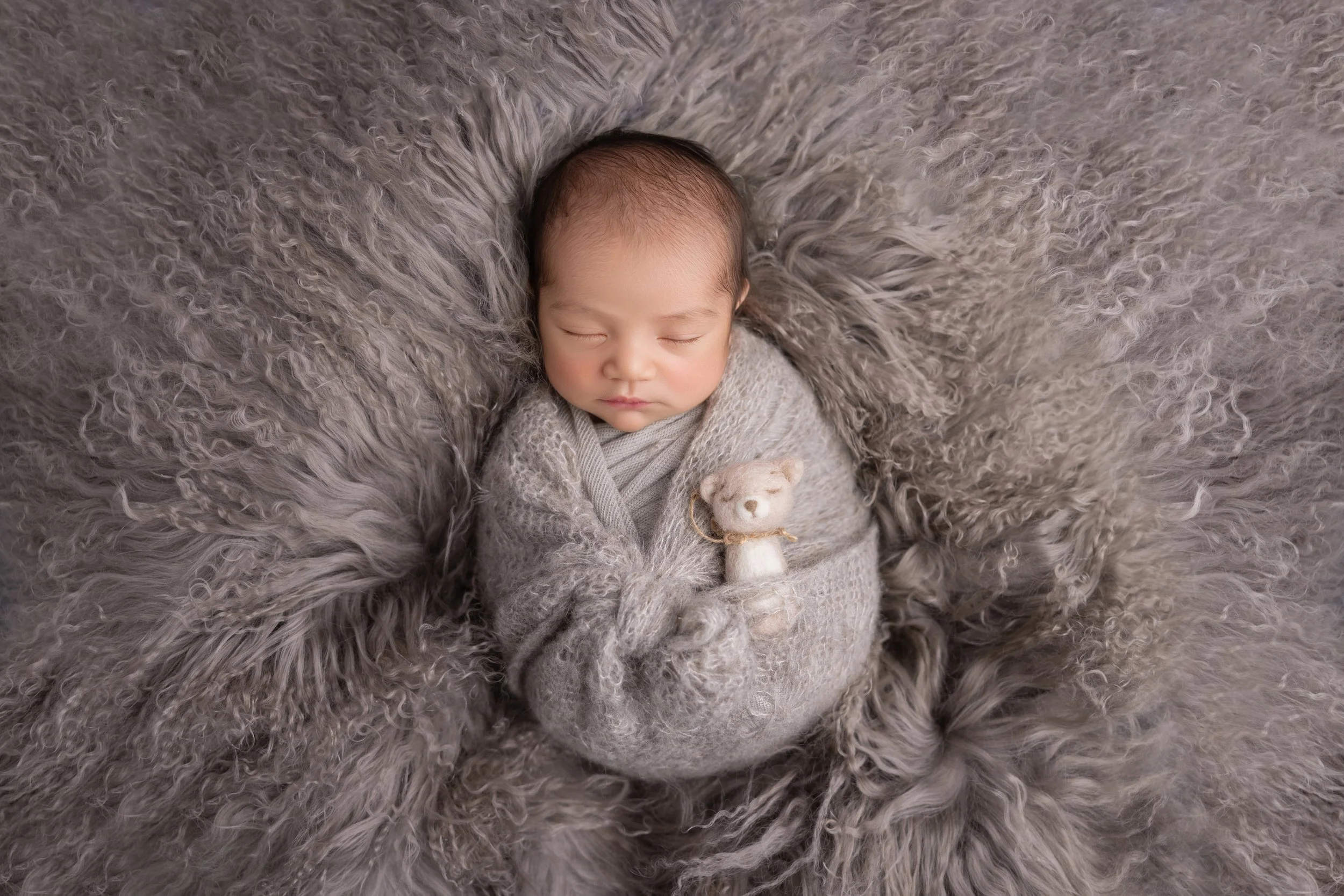 A peacefully sleeping baby wrapped in a grey knitted blanket, lying on a fluffy grey fur blanket, holding a small beige teddy bear.
