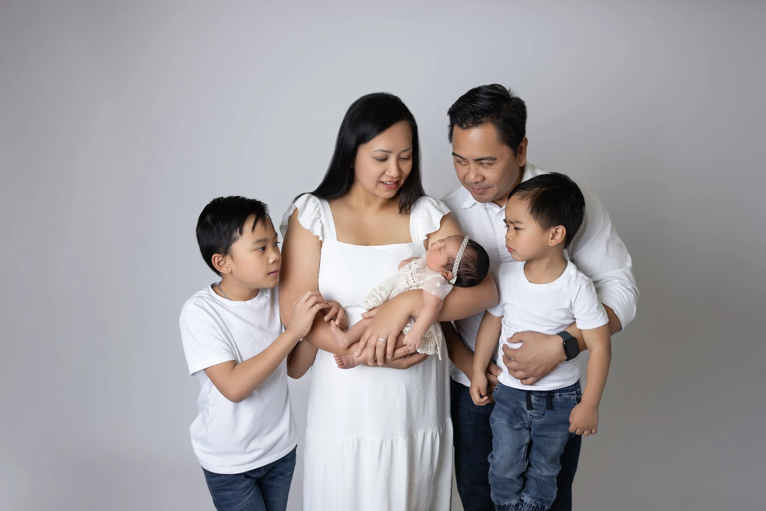 Family of five, including newborn, posing together against a plain background. The mother holds the baby, and the father and two young boys surround them.