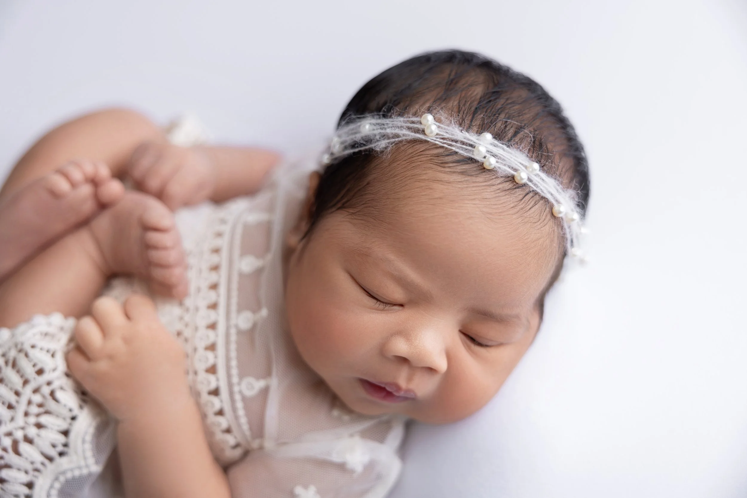 Close-up of a sleeping baby girl with a pearl and white headband, wearing a delicate lace dress, lying on a white surface.