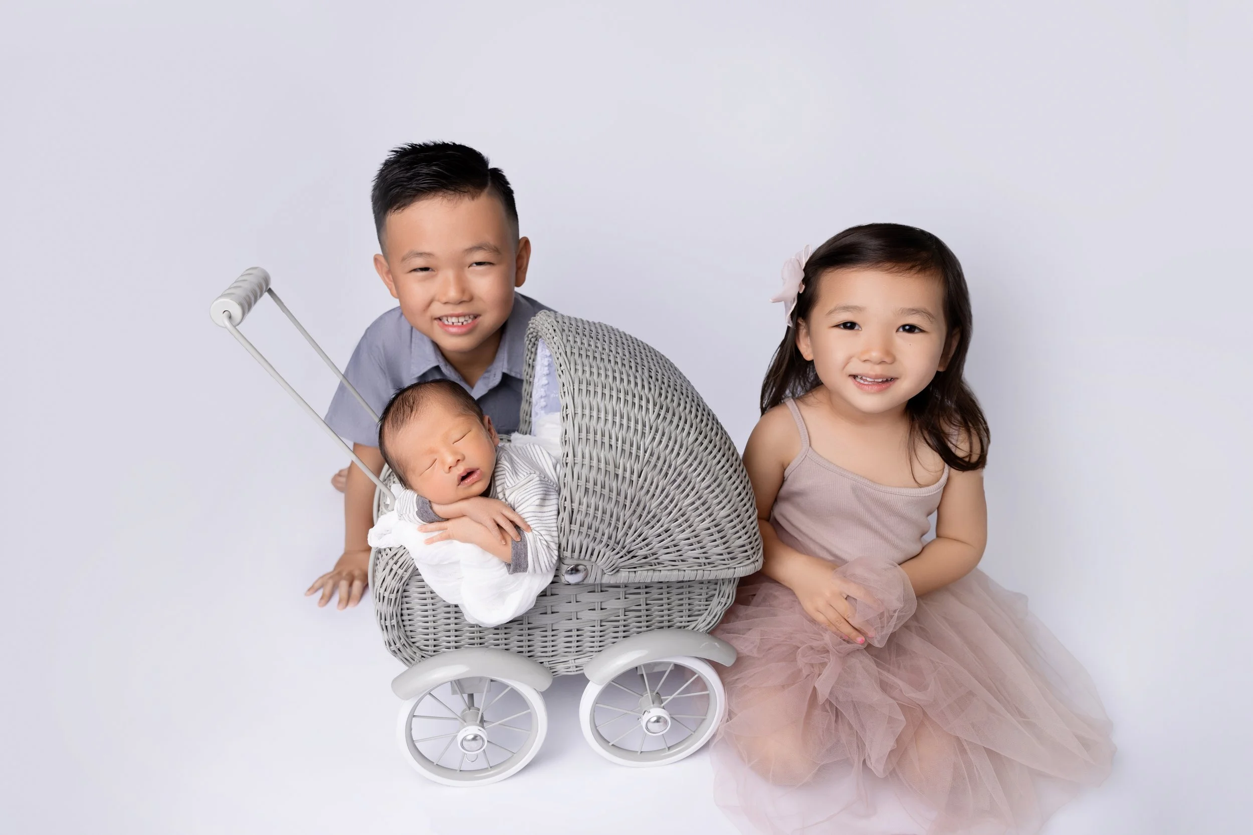 A young boy and girl, with the boy smiling and the girl with a gentle expression, posing with a newborn baby in a white wicker stroller. The girl is sitting beside the stroller wearing a light pink dress with tulle, and her hair is adorned with a sma