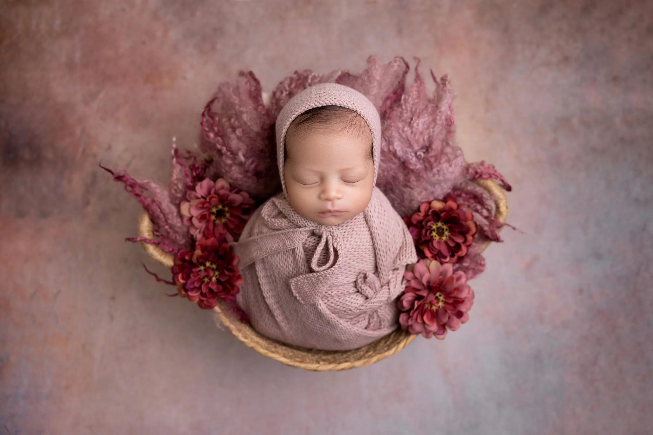 A sleeping baby in a pink knitted outfit and bonnet, laying in a basket with pink and red flowers and a pink textured blanket, on a multicolored background.