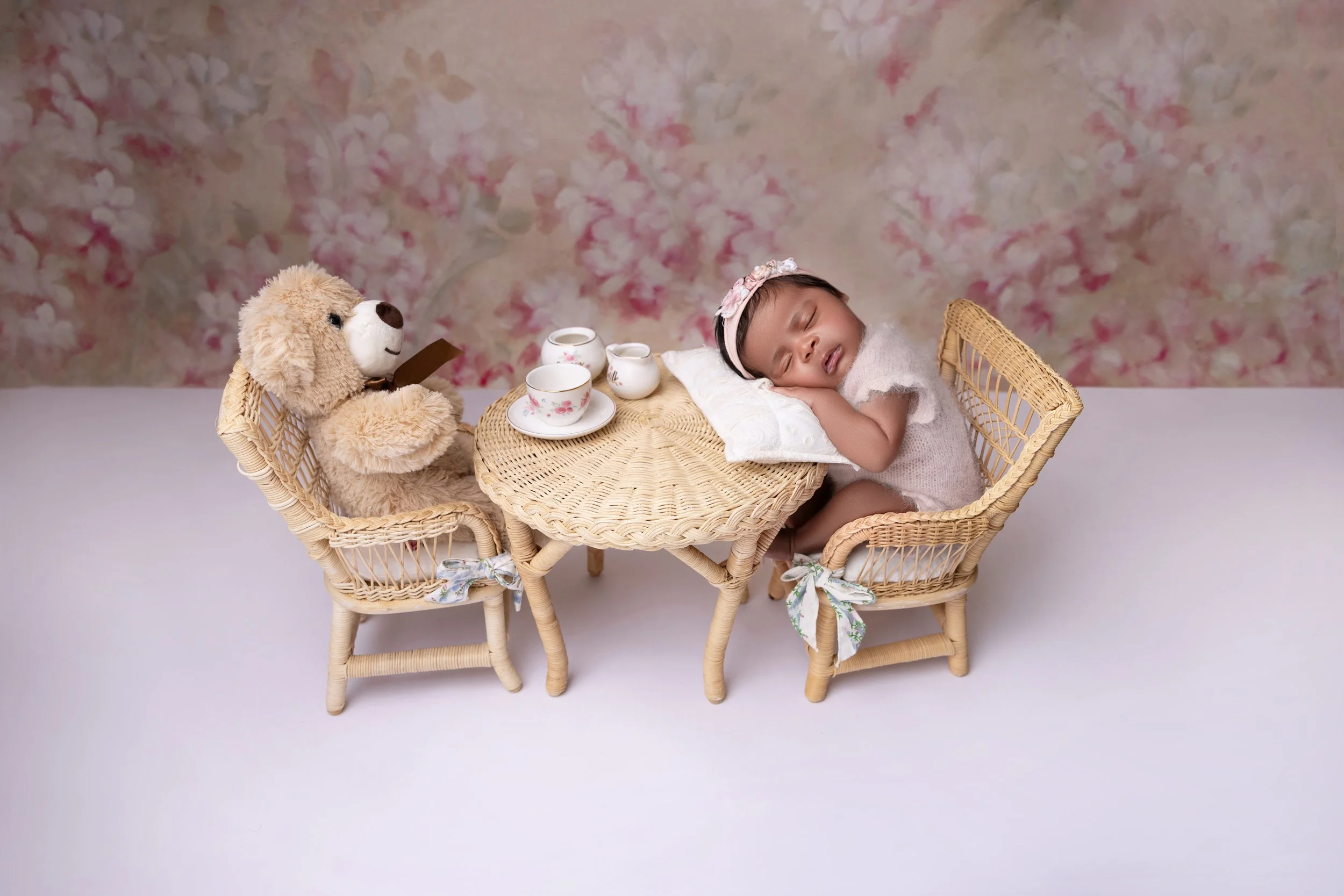 A young child is sleeping with her head resting on a pillow at a small wicker table. She is wearing a light dress and a floral headband. A teddy bear is sitting on a matching wicker chair opposite her, holding a book. The table has a tea set with a t