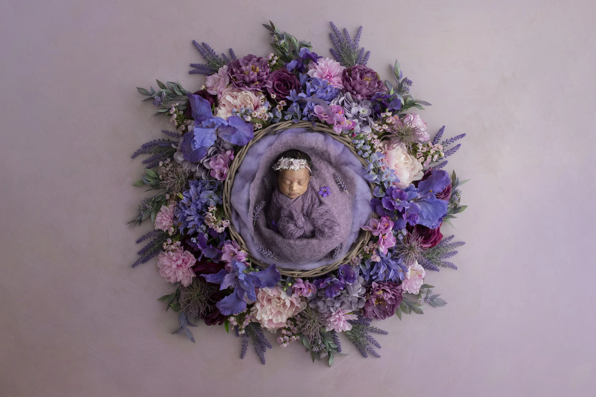 A sleeping baby girl in a purple knitted outfit and floral headband, wrapped in purple blanket, lying inside a round wicker basket surrounded by an arrangement of purple, pink, and lavender flowers and greenery on a light pink background.
