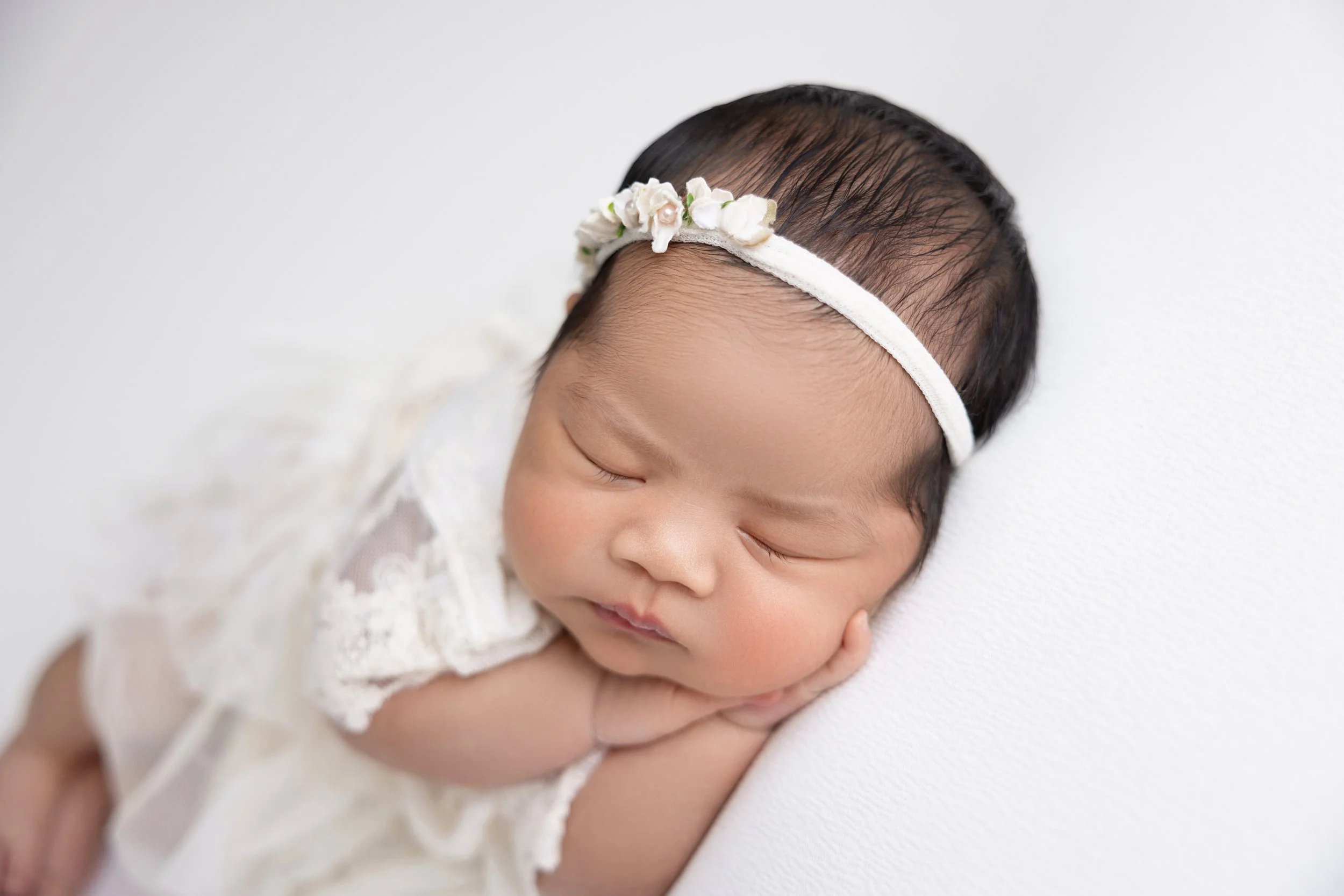 Close-up of a sleeping baby girl with a floral headband, resting her head on her hands, dressed in a white lace outfit on a white surface.