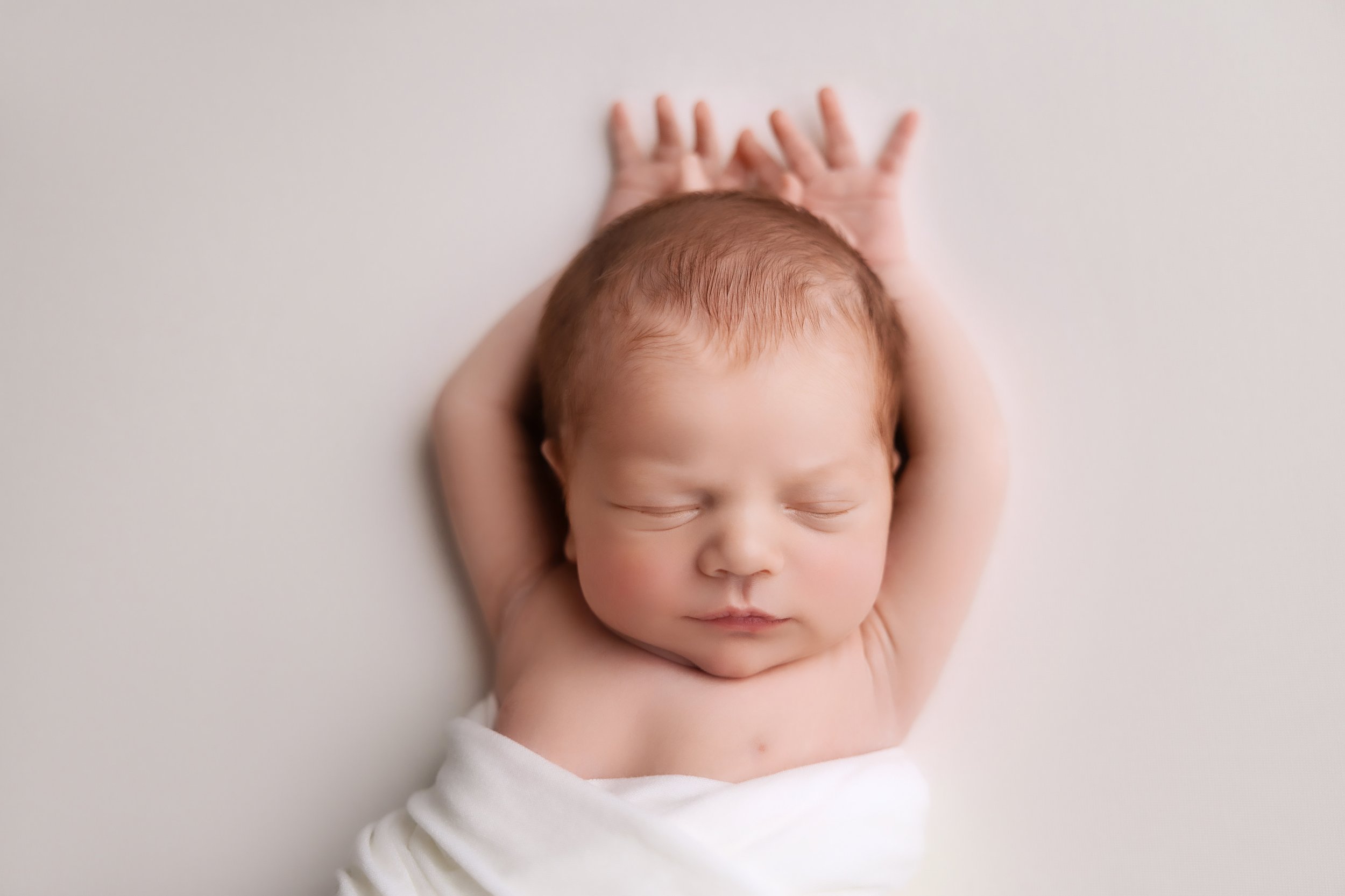 A sleeping newborn baby with arms stretched above the head, wrapped in a white cloth, lying on a soft white surface.