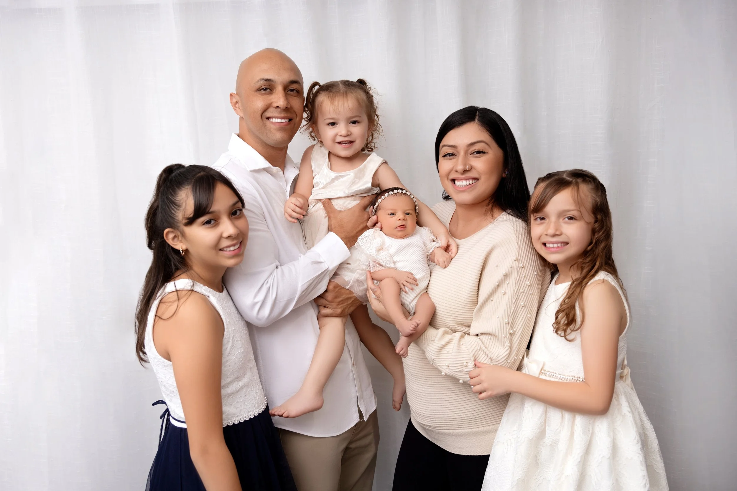 Family portrait of a man, woman, and four girls standing together smiling against a white background, with the man holding a young girl and the woman holding a baby.