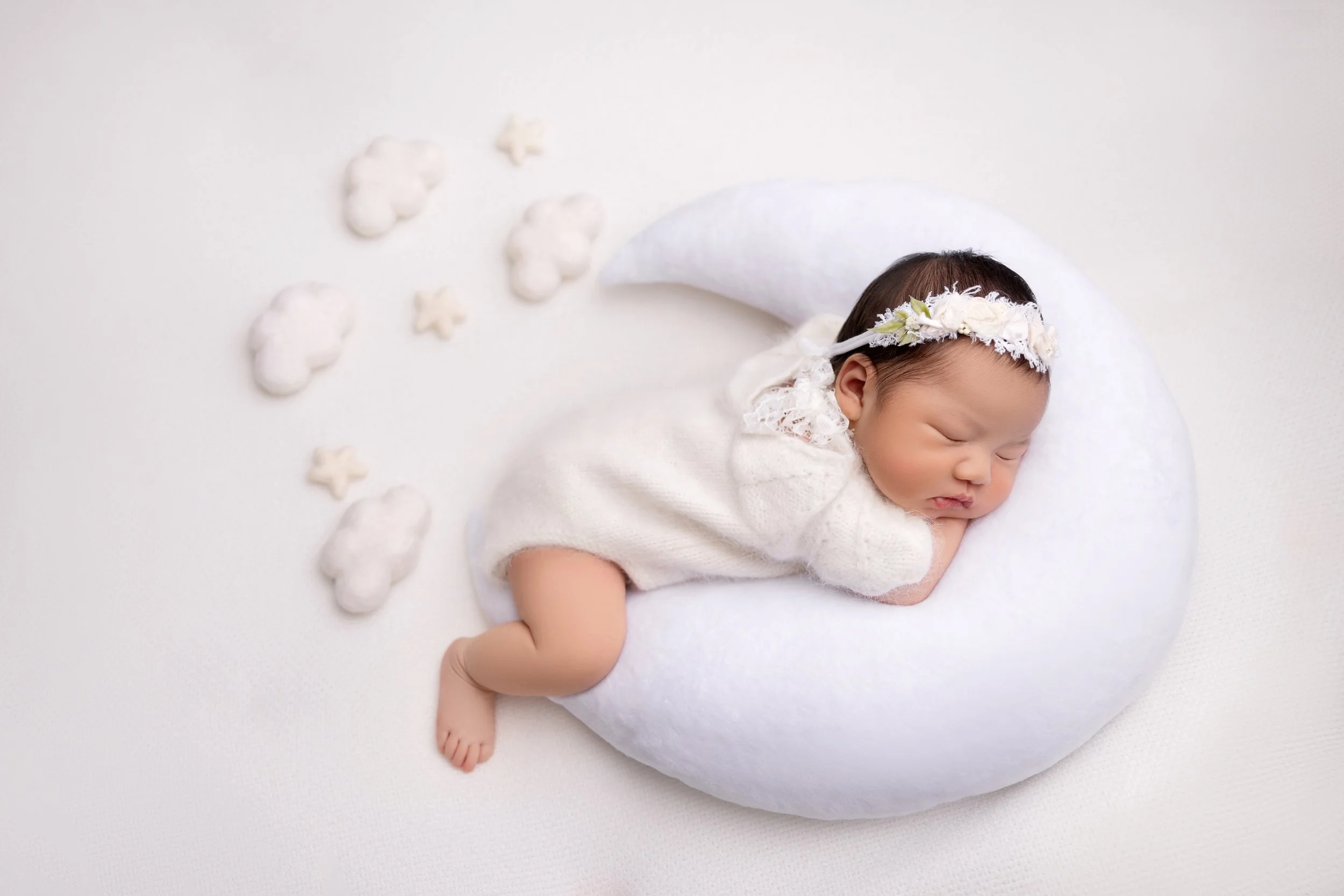 A peacefully sleeping newborn baby girl, lying on her side on a white crescent moon-shaped pillow, wearing a white knit outfit and a floral headband, with soft cloud and star-shaped decorations nearby.