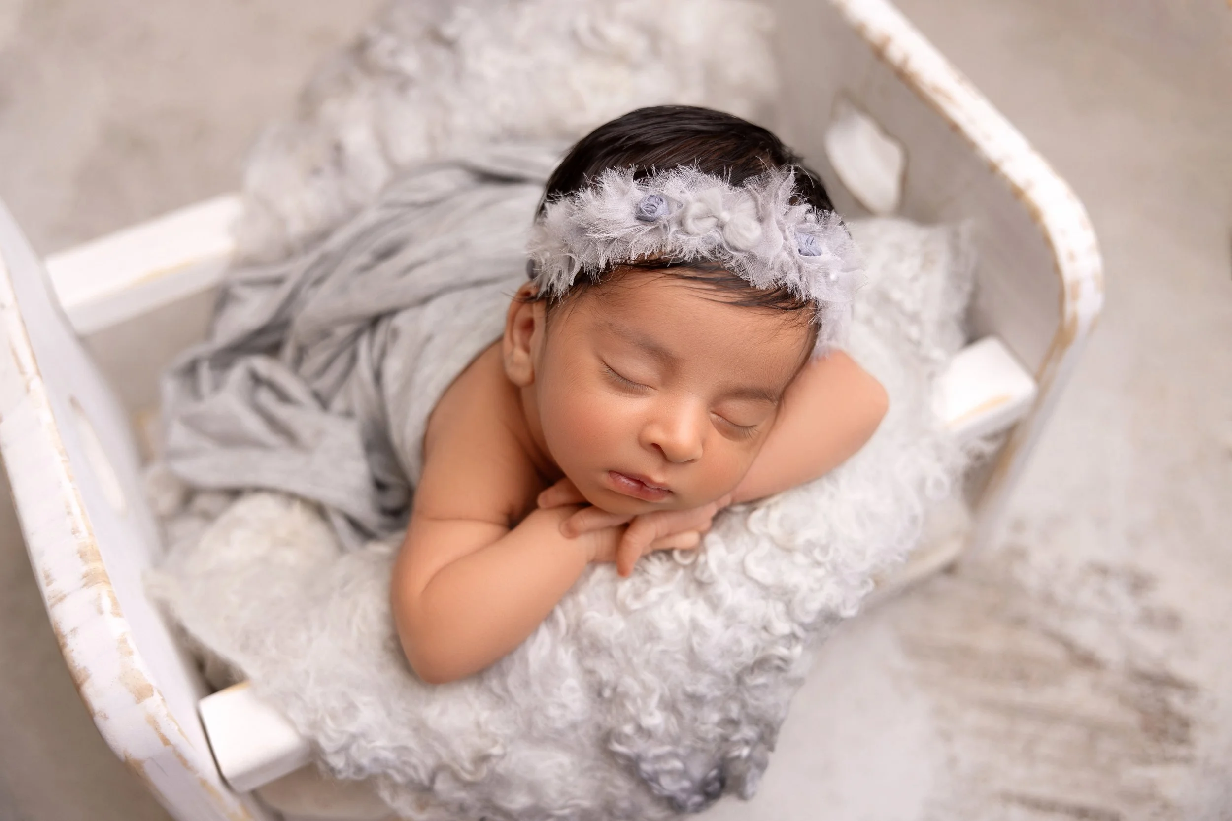 A sleeping baby girl with a floral headband, lying on her side in a small white bed with soft bedding.