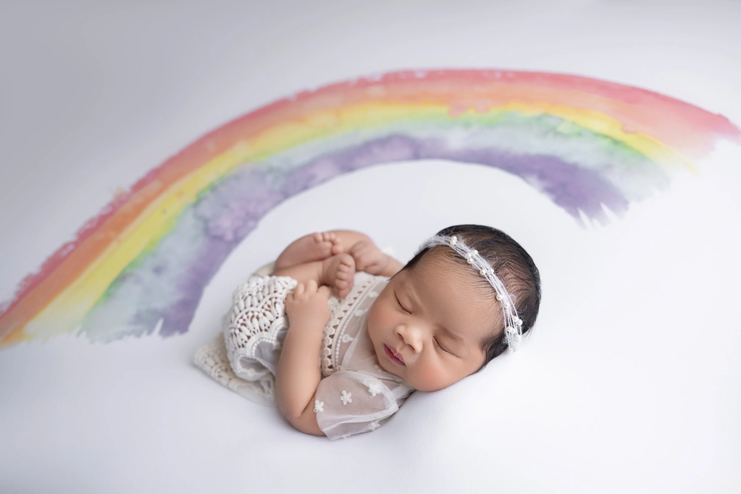 Sleeping baby girl with a pearl headband, dressed in lace clothing, lying on white surface with a painted rainbow above her.