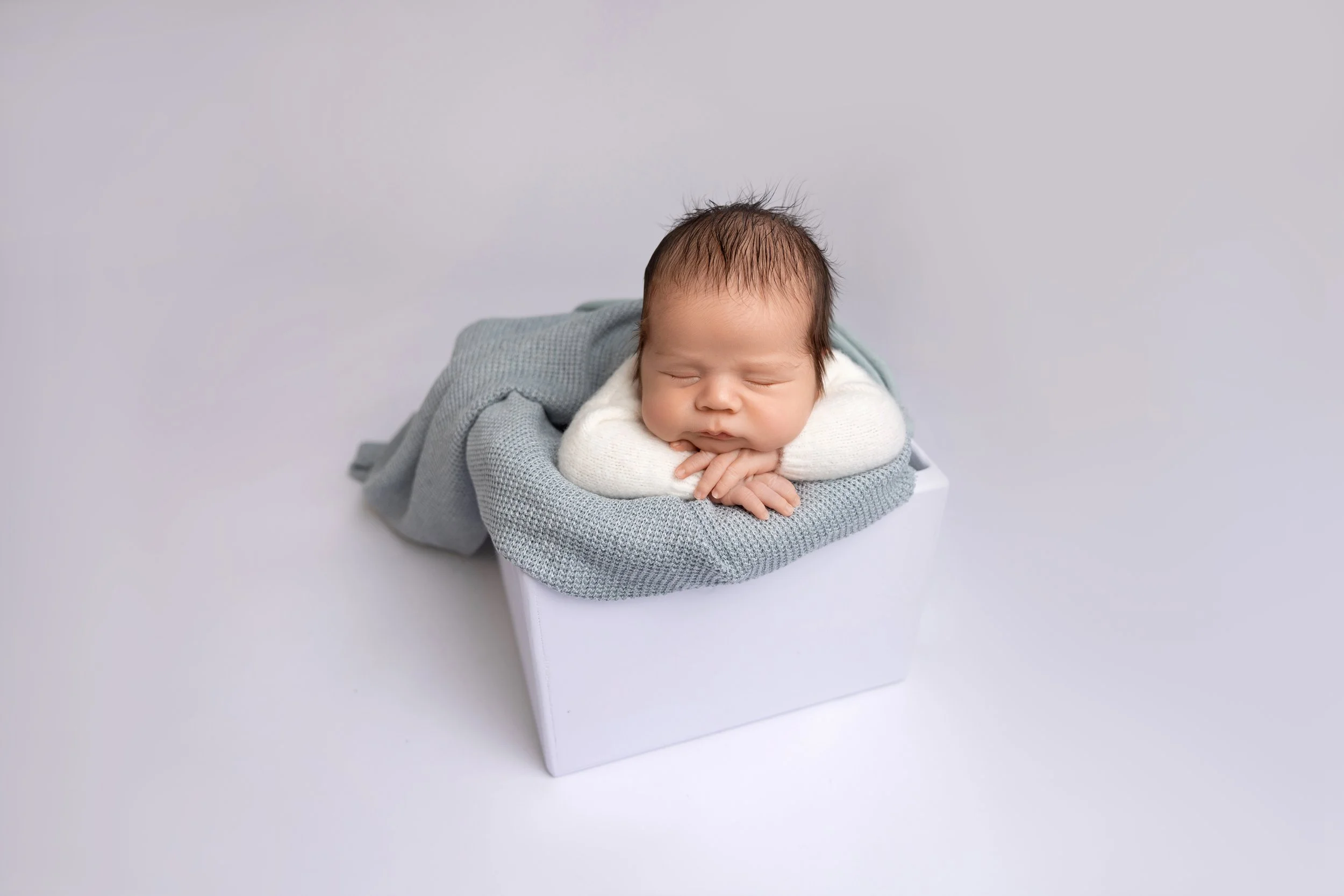 A sleeping baby with dark hair lying on a gray knitted blanket inside a white box, resting their head on folded hands.
