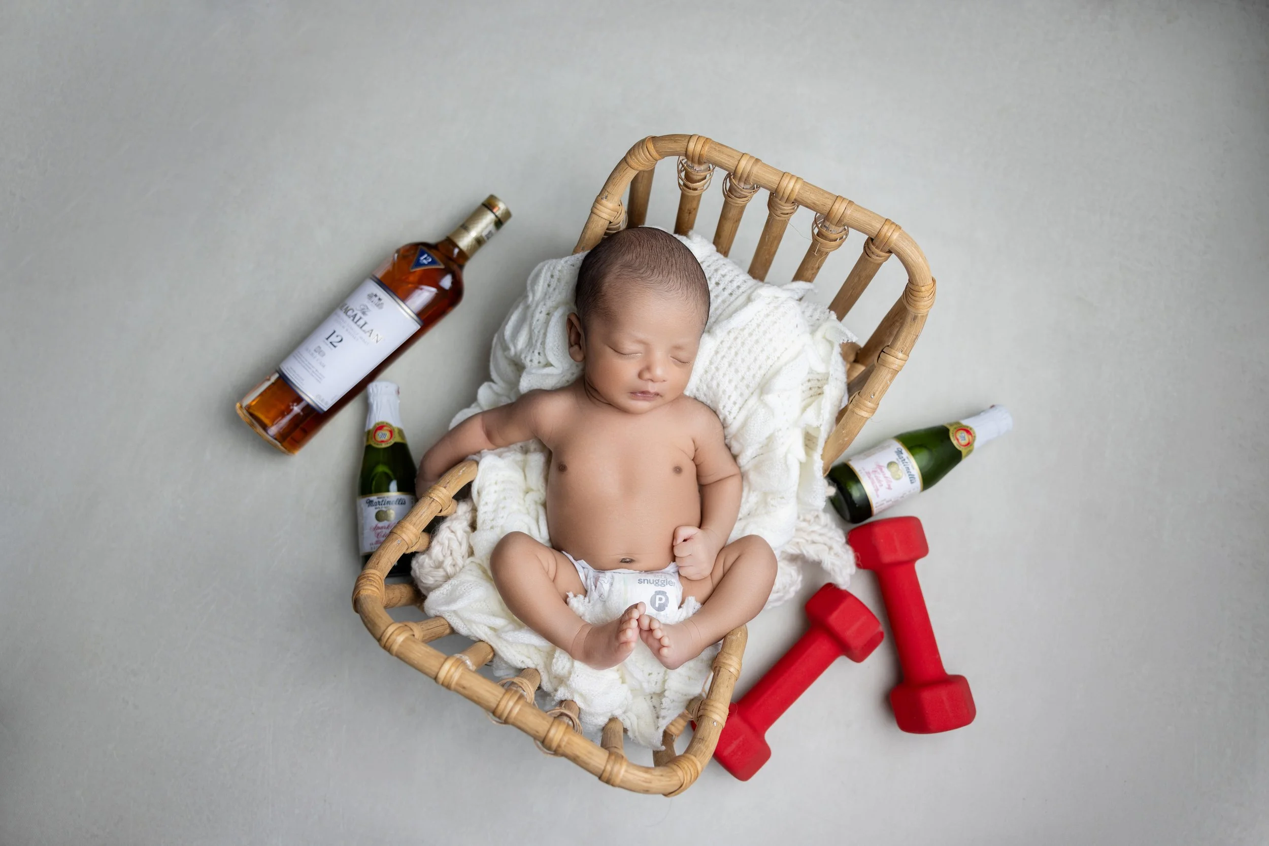 A baby sleeping in a wicker basket, surrounded by miniature bottles of alcohol, two small red dumbbells, with a neutral background.