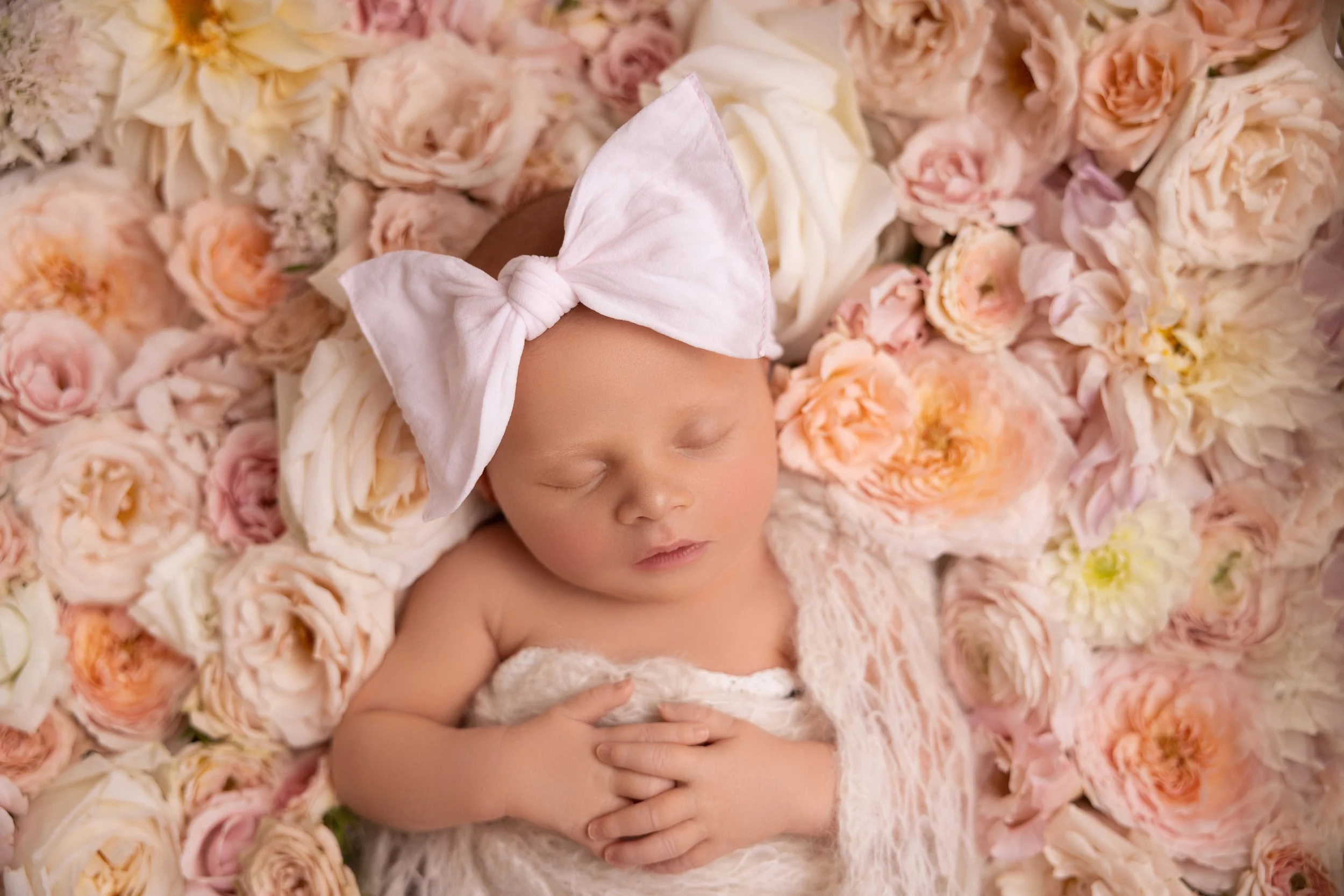 A sleeping newborn baby girl wearing a large pink bow headband, surrounded by a bed of soft pastel-colored roses and peonies.
