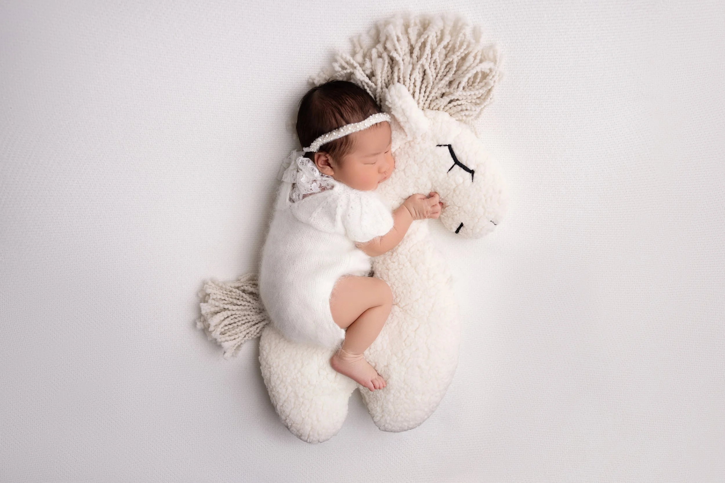 A sleeping baby in white clothes, curled on a plush unicorn pillow on a plain white background.