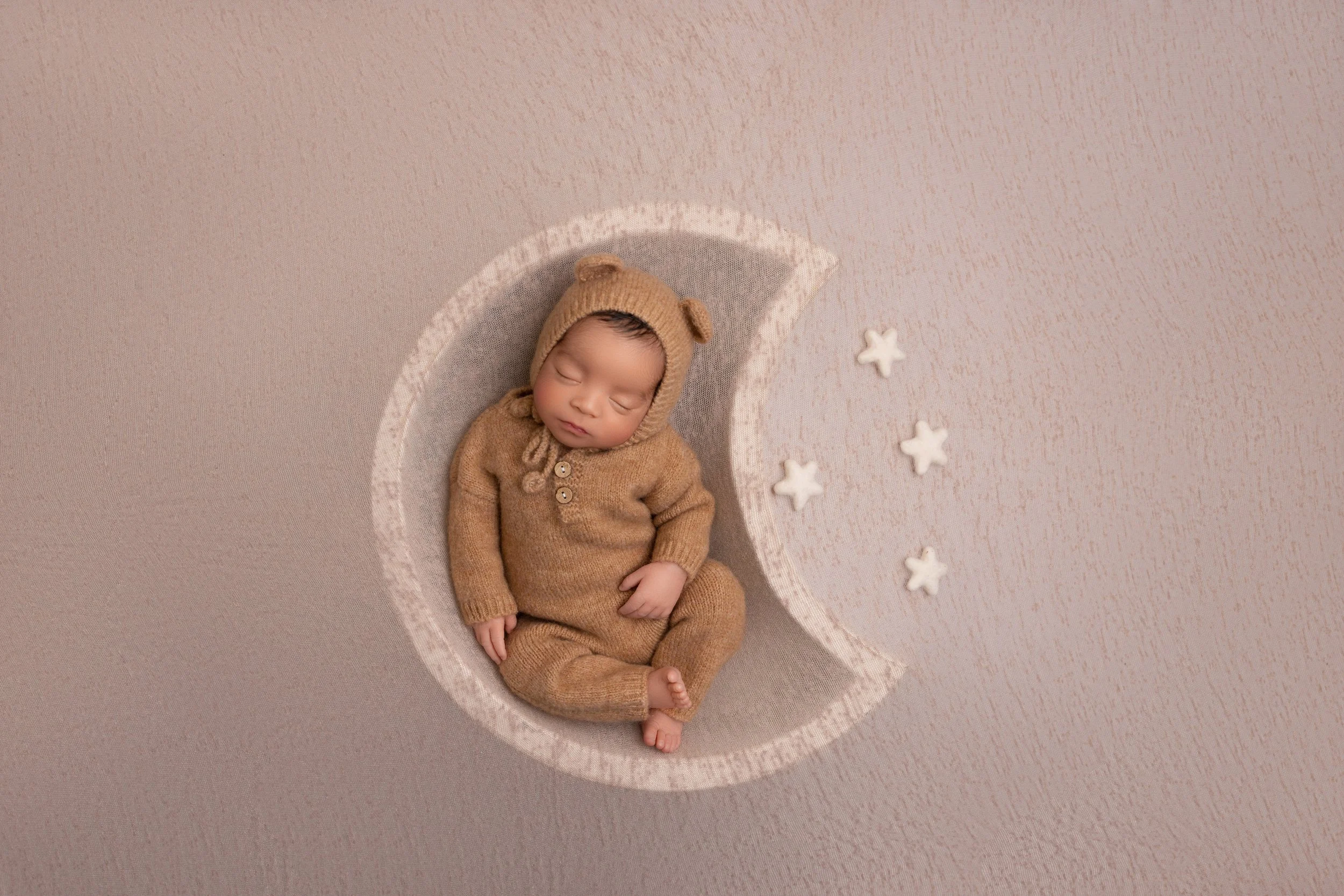 A sleeping baby dressed in a brown bear onesie and hat, lying in a crescent moon-shaped bed with white star decorations on a beige background.