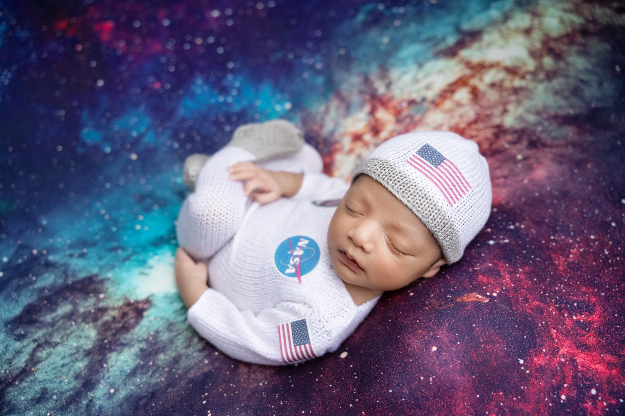 A sleeping baby dressed in a NASA-themed outfit with an American flag hat, lying on a colorful galaxy-themed blanket.