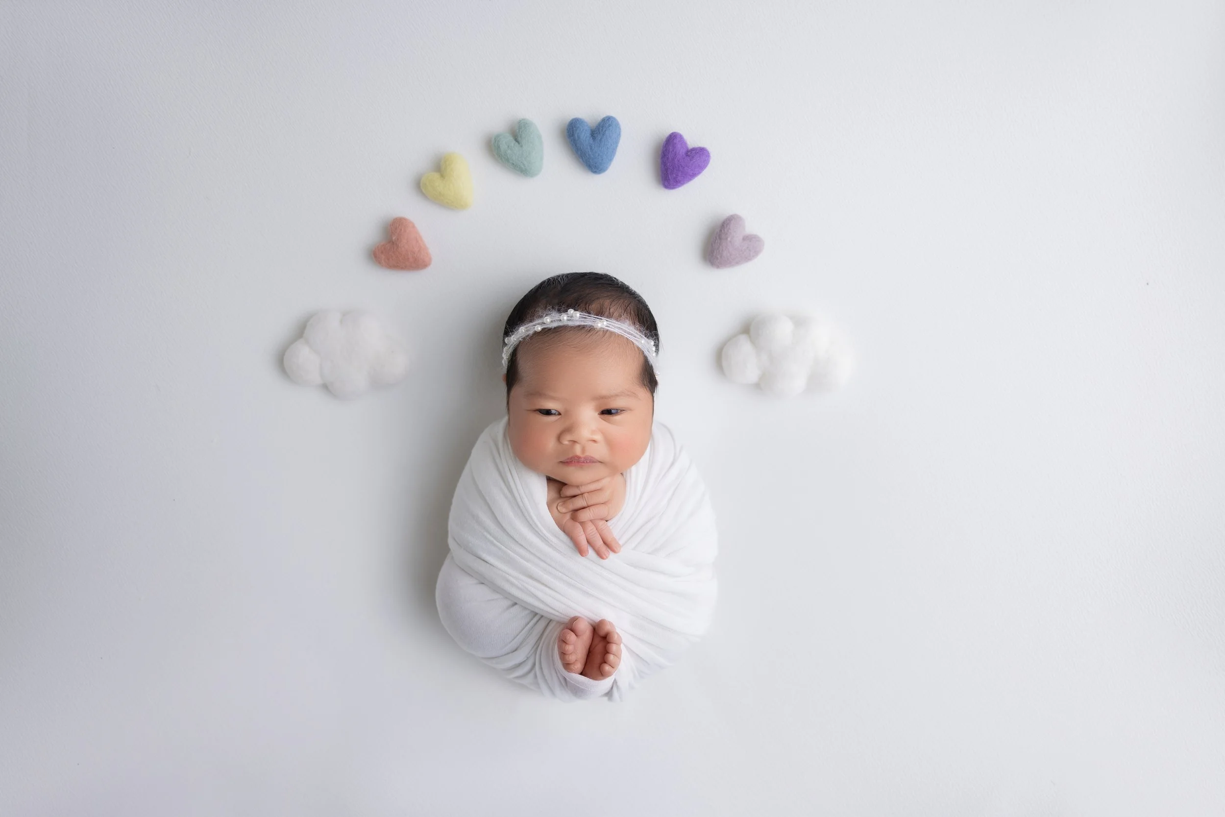 A baby wrapped in a white blanket, lying on a white surface, surrounded by colorful felt hearts and clouds, creating a cloud-like scene.