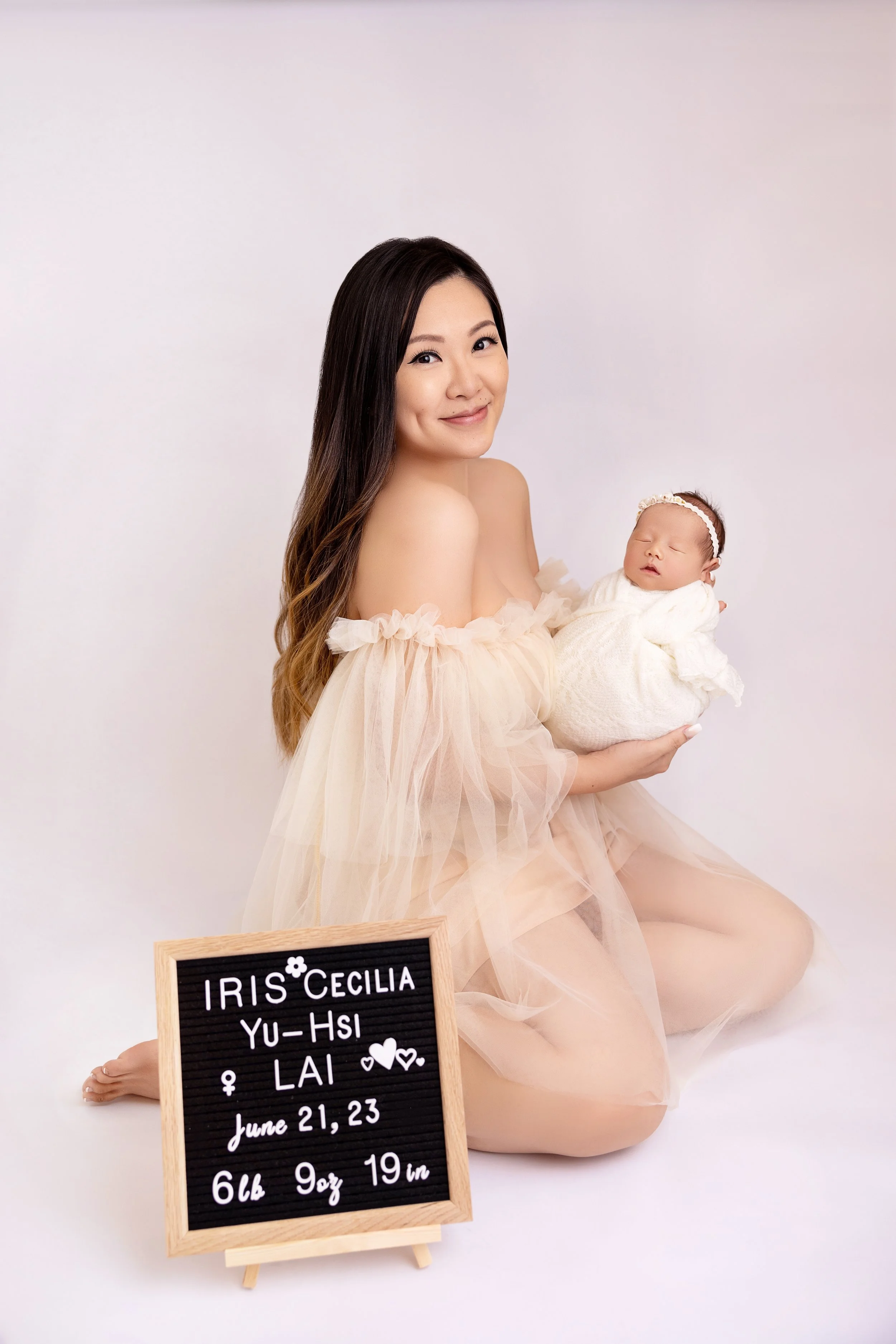 A woman with long brown hair kneeling on the floor, holding a sleeping baby wrapped in a white blanket, against a plain pinkish-white background. There is a small chalkboard sign in front of them with information about the baby, including name, birth