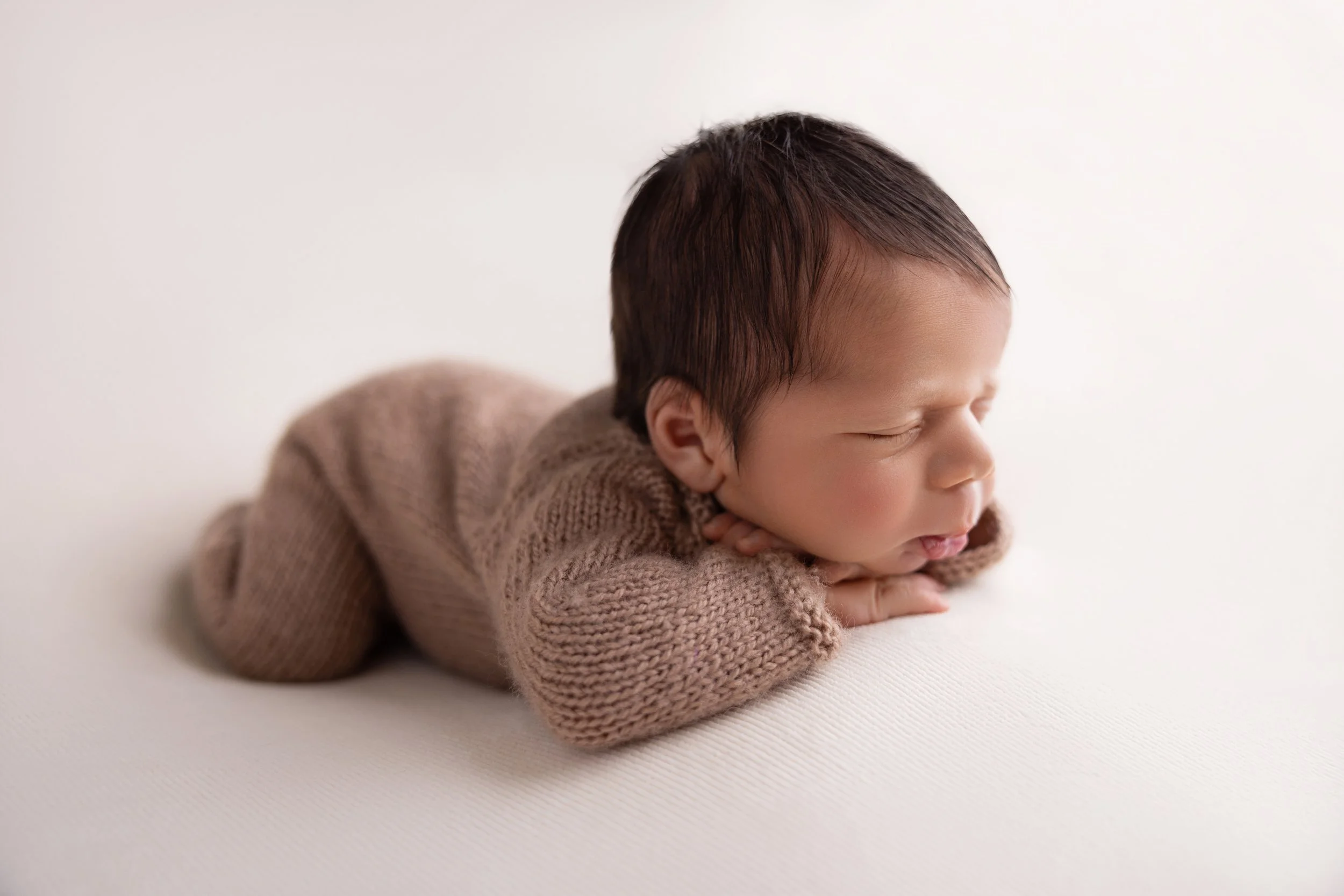 A newborn baby sleeping on a white surface, wearing a brown knitted outfit.