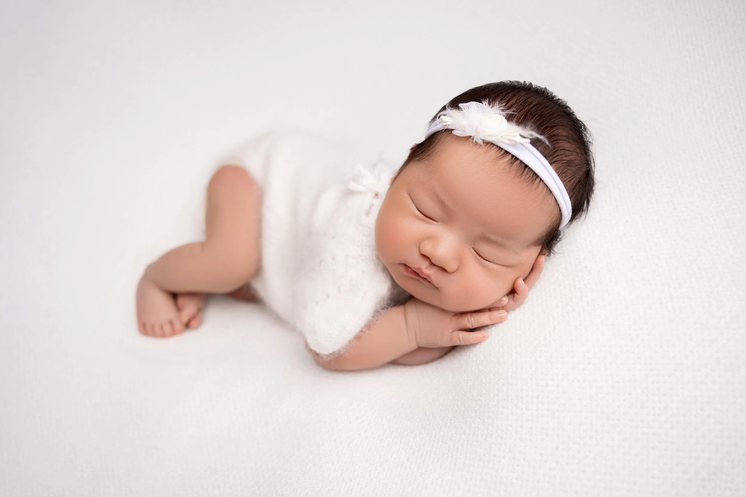A sleeping baby girl lying on her side with her head resting on her hands, dressed in white with a white headband, on a white textured surface.