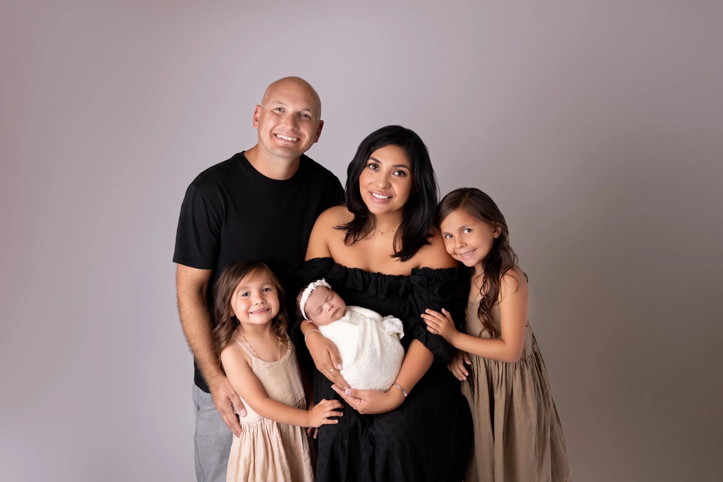 Family portrait of five, including a woman holding a sleeping baby, a man, and three young girls, all smiling and dressed casually, against a plain gray background.