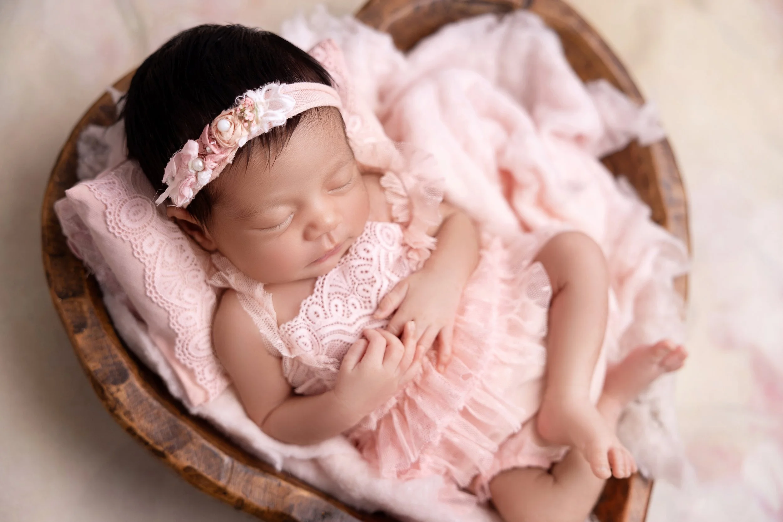 A sleeping baby girl with a headband with flowers, lying in a wooden basket on a soft pink blanket.