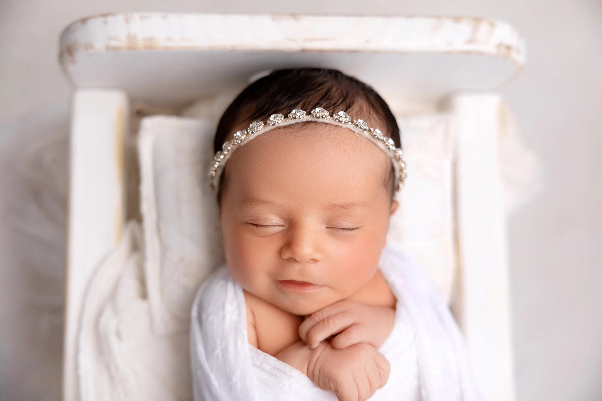 A sleeping baby girl with a jeweled headband, wrapped in a white blanket, lying on a small white bed.