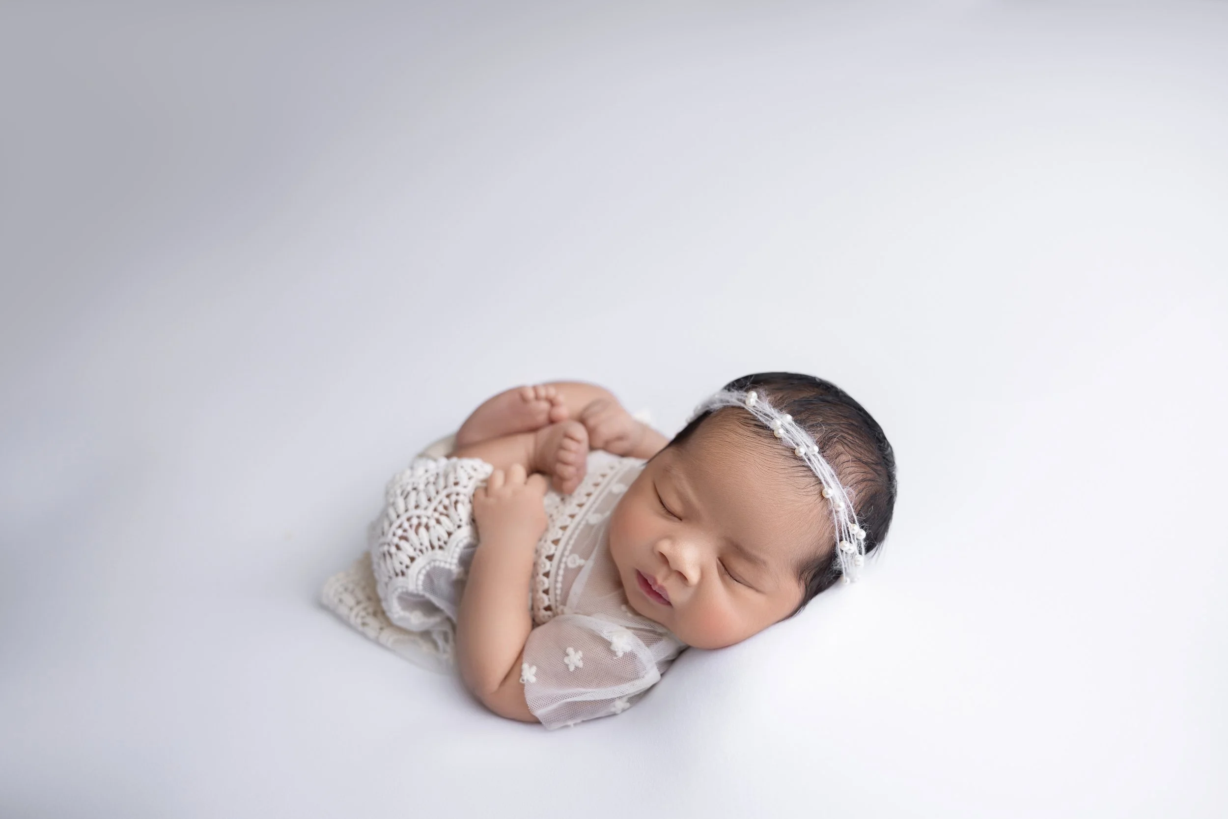 A sleeping newborn baby girl lying on a white surface, dressed in a lace dress and wearing a pearl headband.