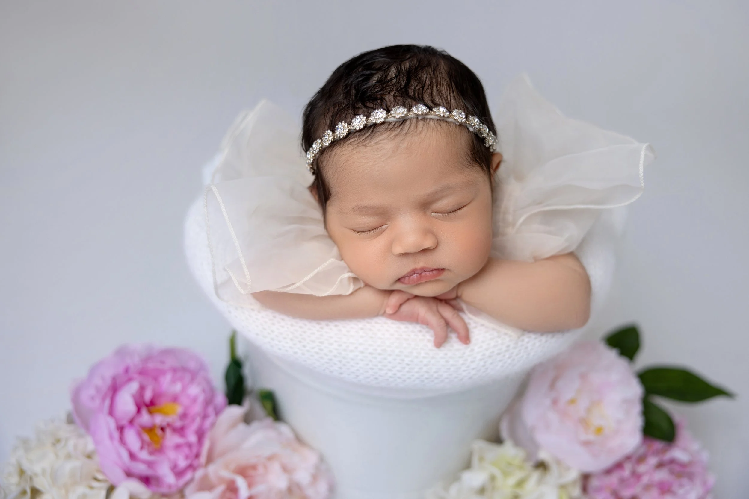 Sleeping baby girl with dark hair and a jeweled headband, resting her chin on her arms, surrounded by pink and white flowers.